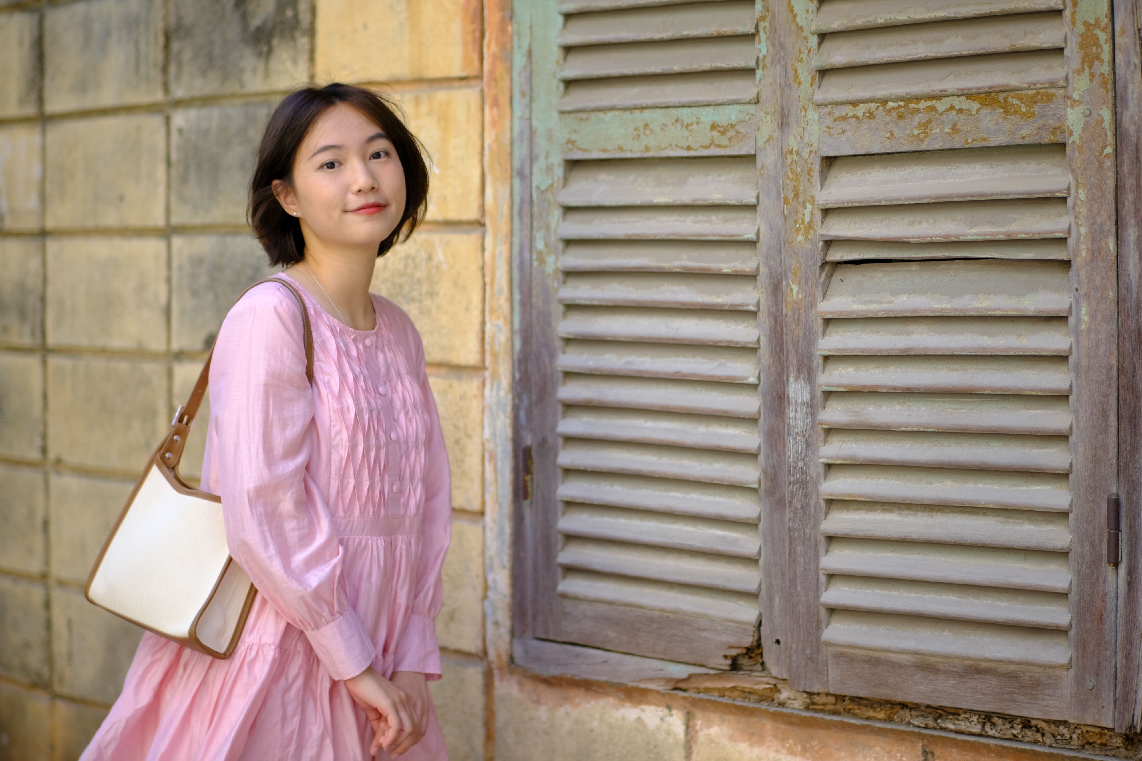 Young woman poses gracefully in a pink dress by a rustic window in Ho Chi Minh City.