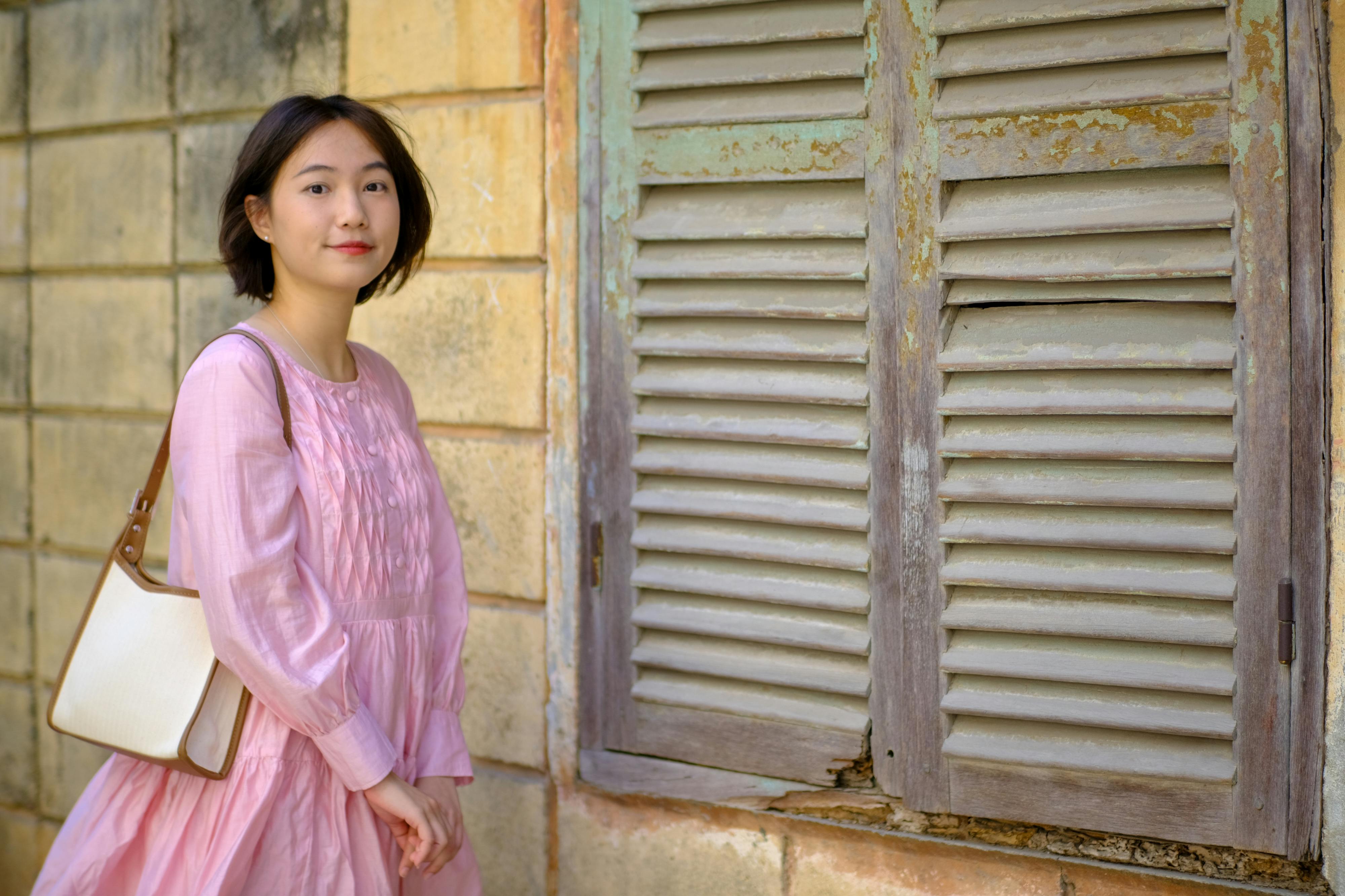 ฟรี ภาพถ่ายอันสงบเงียบของหญิงสาวในชุดสีชมพูข้างหน้าต่างเก่าแก่ในเมืองโฮจิมินห์ ประเทศเวียดนาม คลังภาพถ่าย