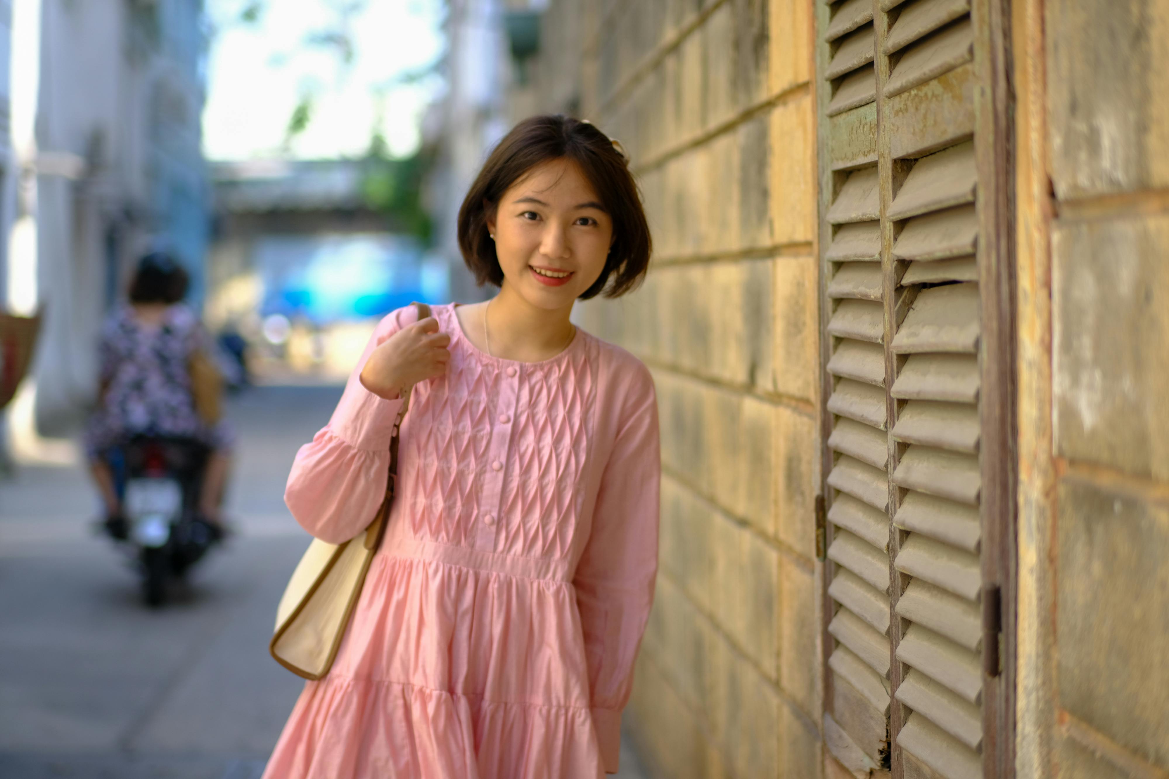 Miễn phí Một cô gái trẻ tươi cười trong chiếc váy hồng tạo dáng trước bức tường mộc mạc ở Thành phố Hồ Chí Minh. Ảnh lưu trữ