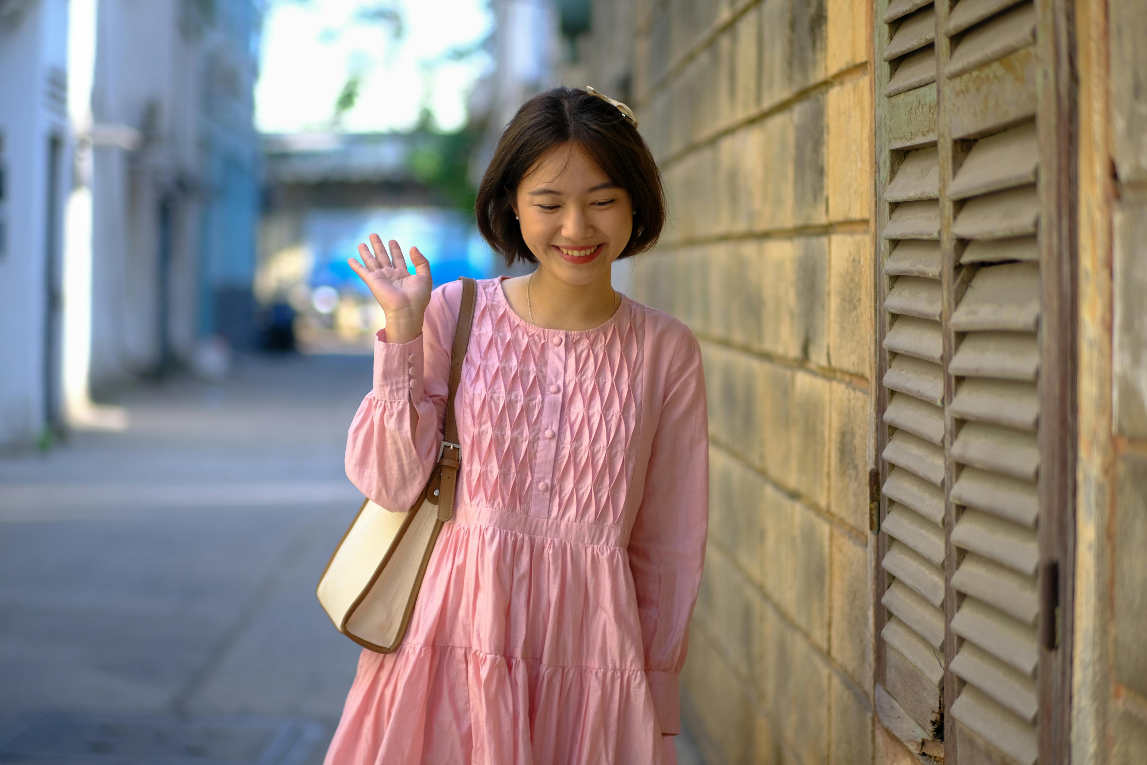 ingyenes Egy rózsaszín ruhás fiatal nő vidáman integet, miközben végigsétál Ho Si Minh-város egyik sikátorában. Stockfotó