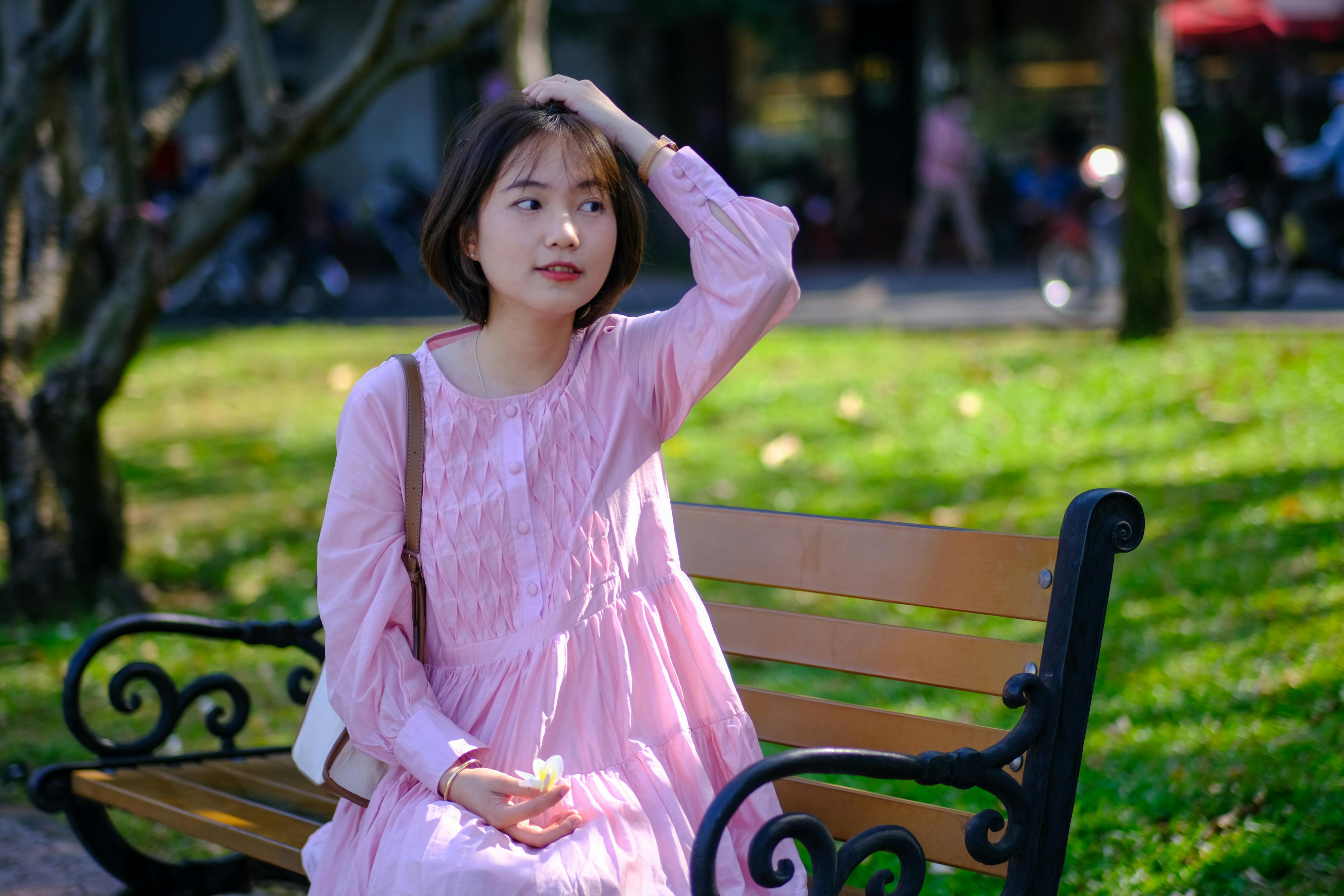 免費 一位身穿粉紅色洋裝的年輕女子坐在胡志明市陽光明媚的公園長椅上。 圖庫相片