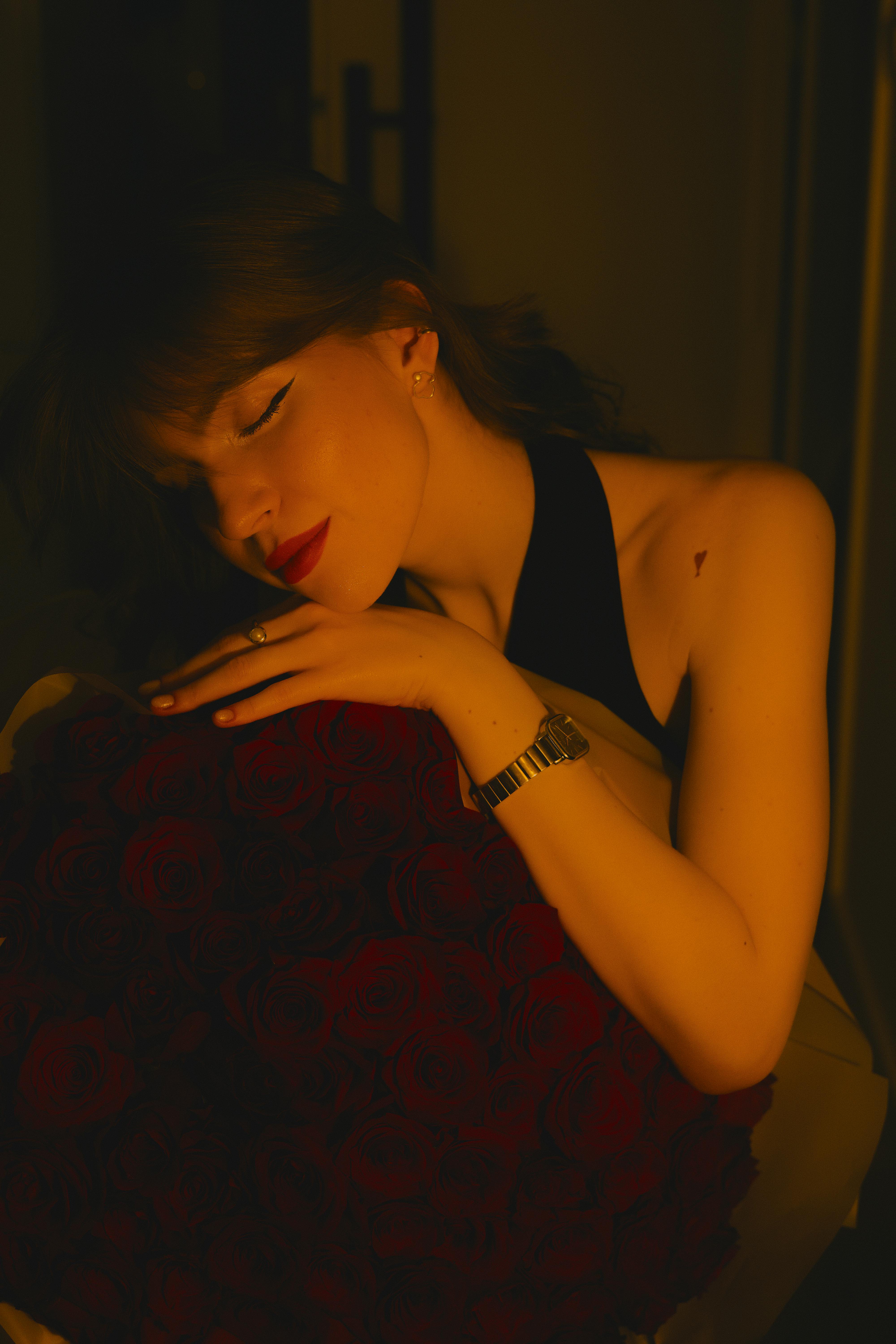 免費 一位神態安詳的女子在溫暖柔和的燈光下抱著一束紅玫瑰，散發著浪漫與寧靜的氣息。 圖庫相片