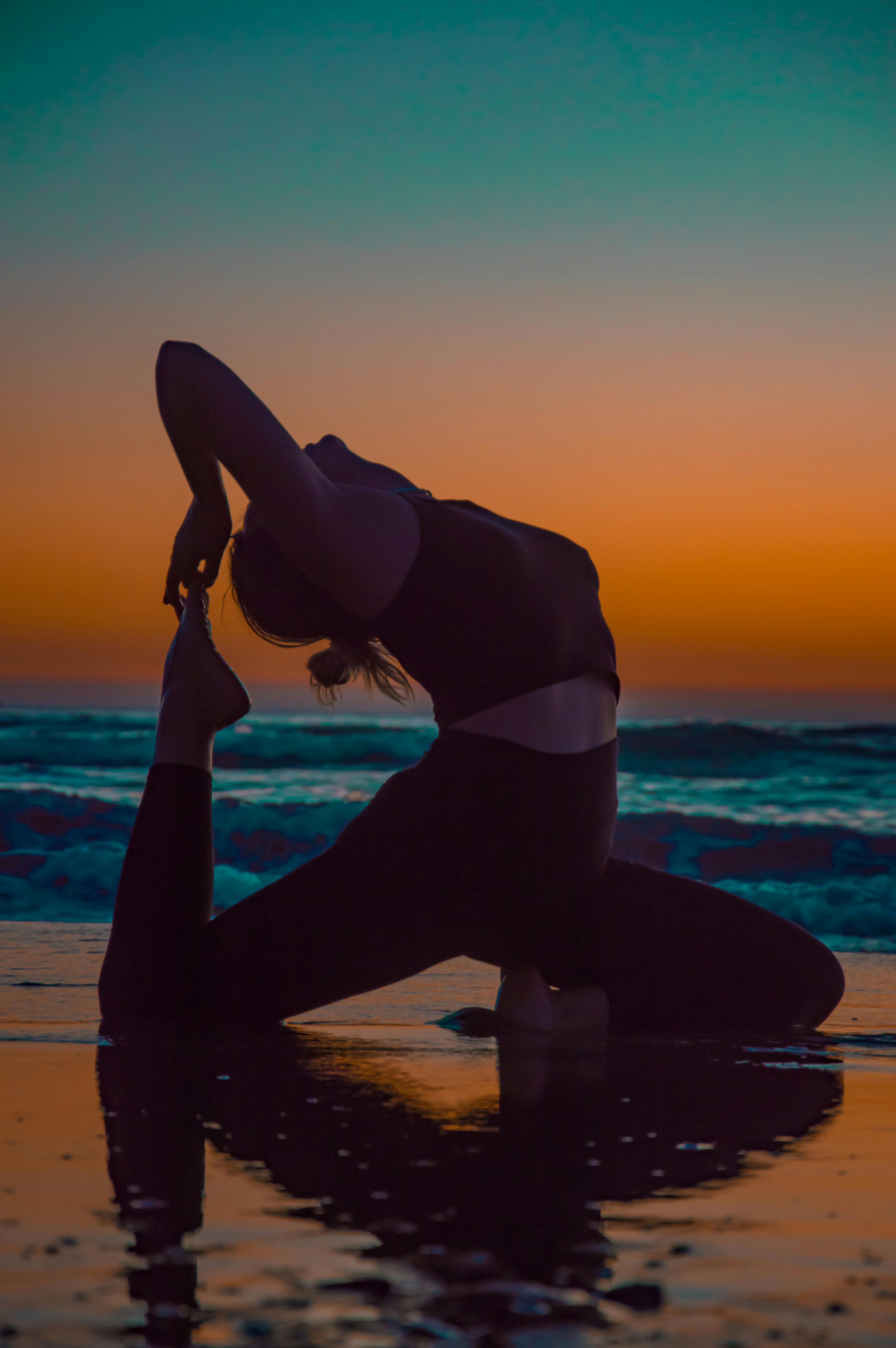 Woman doing yoga at sunset on the beach with teal ocean and orange sky - active recovery and mobility workout