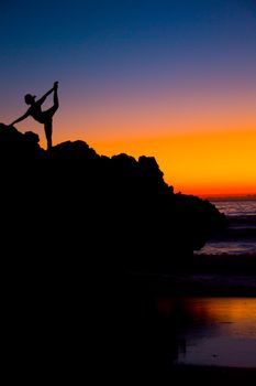Silhouetted yoga pose at sunset on coastal rocks with vibrant sky.