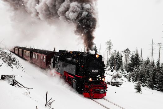 A classic steam train travels through a snowy forest, emitting thick smoke on a winter day.