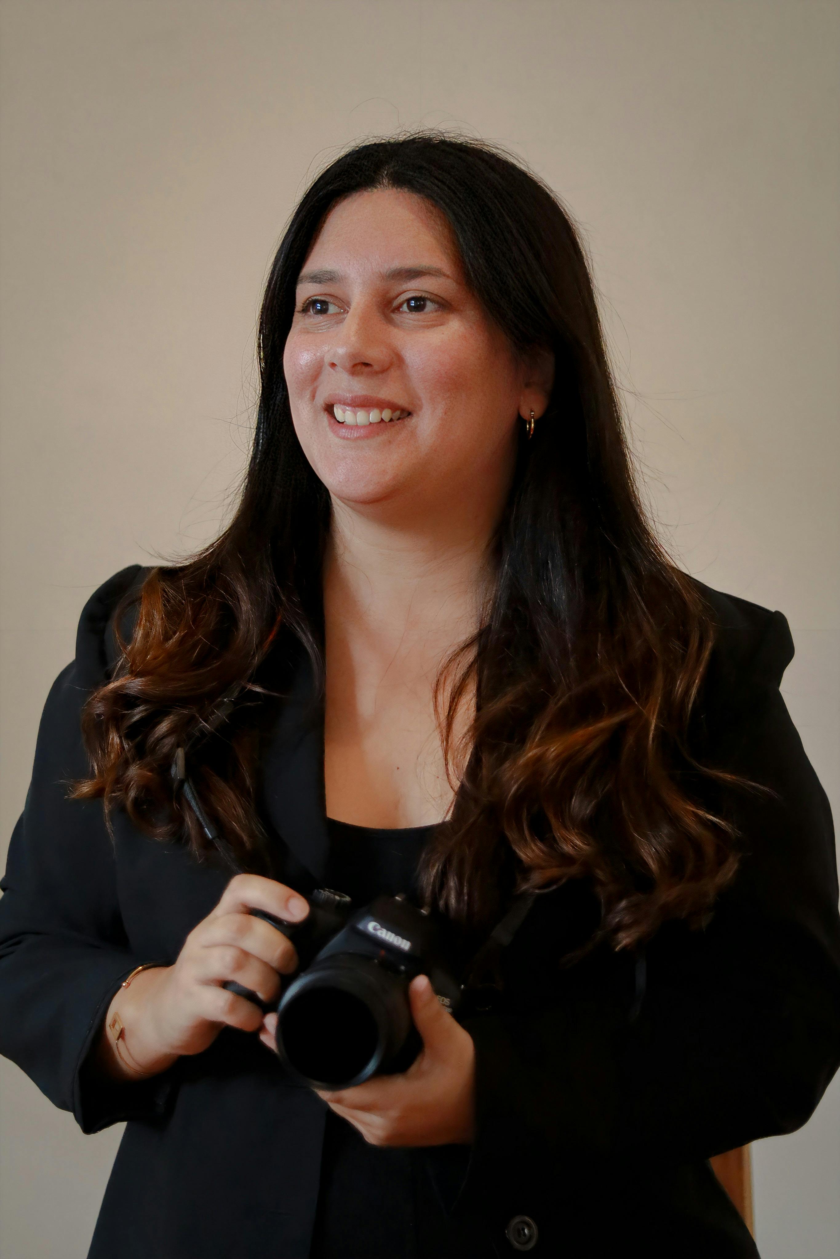 免費 一位面帶微笑的女子手持相機，身穿黑色套裝。室內肖像照。 圖庫相片