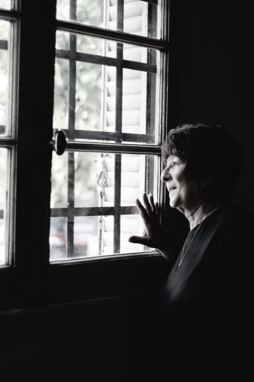 Free Senior woman gazing thoughtfully out of a window, embracing natural light indoors. Stock Photo