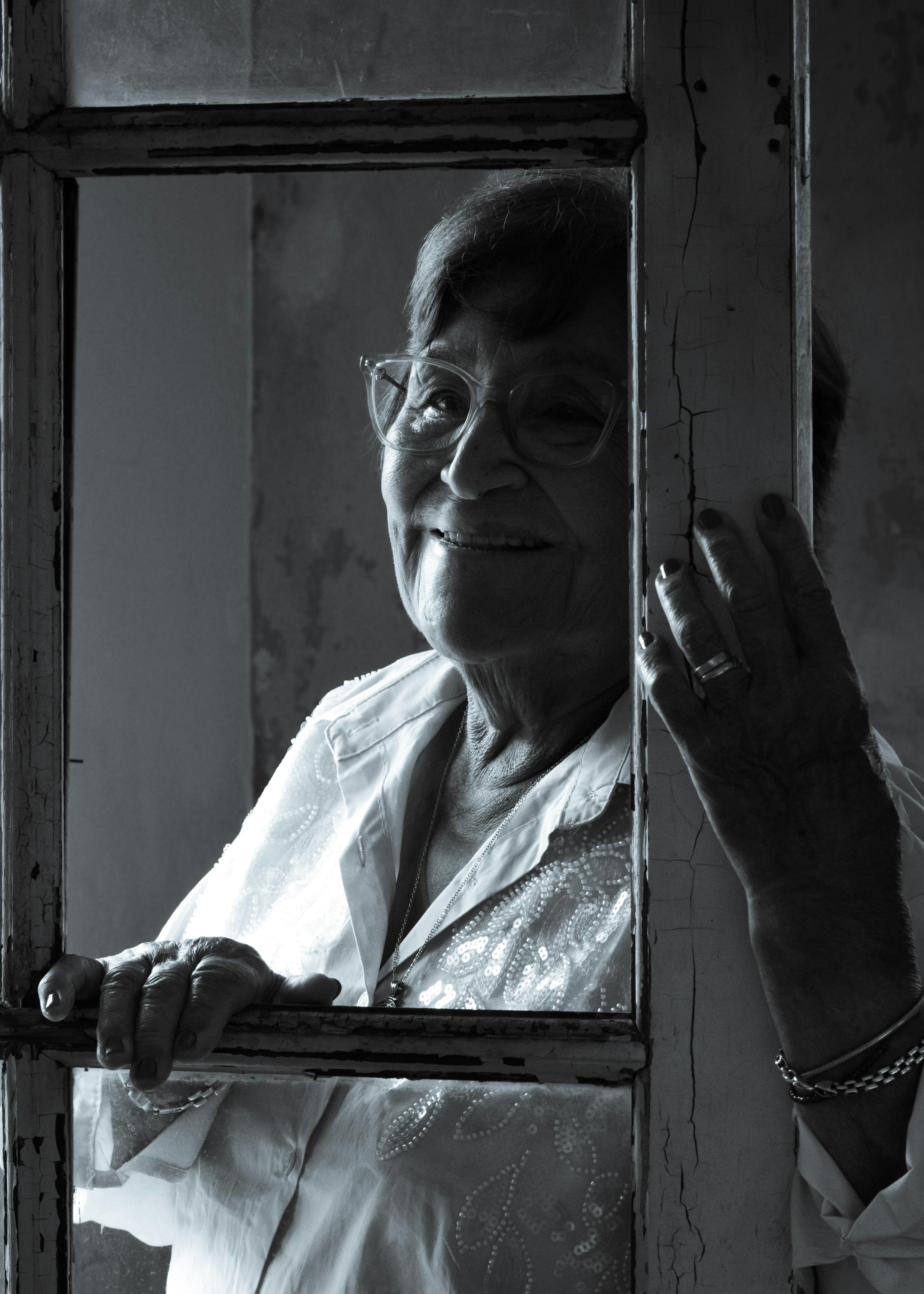 Free Portrait of a senior woman with glasses smiling through a window, invoking nostalgia and warmth. Stock Photo
