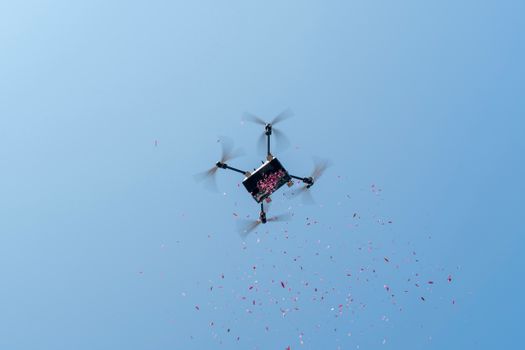 A drone disperses pink petals against a clear blue sky, captured from below.