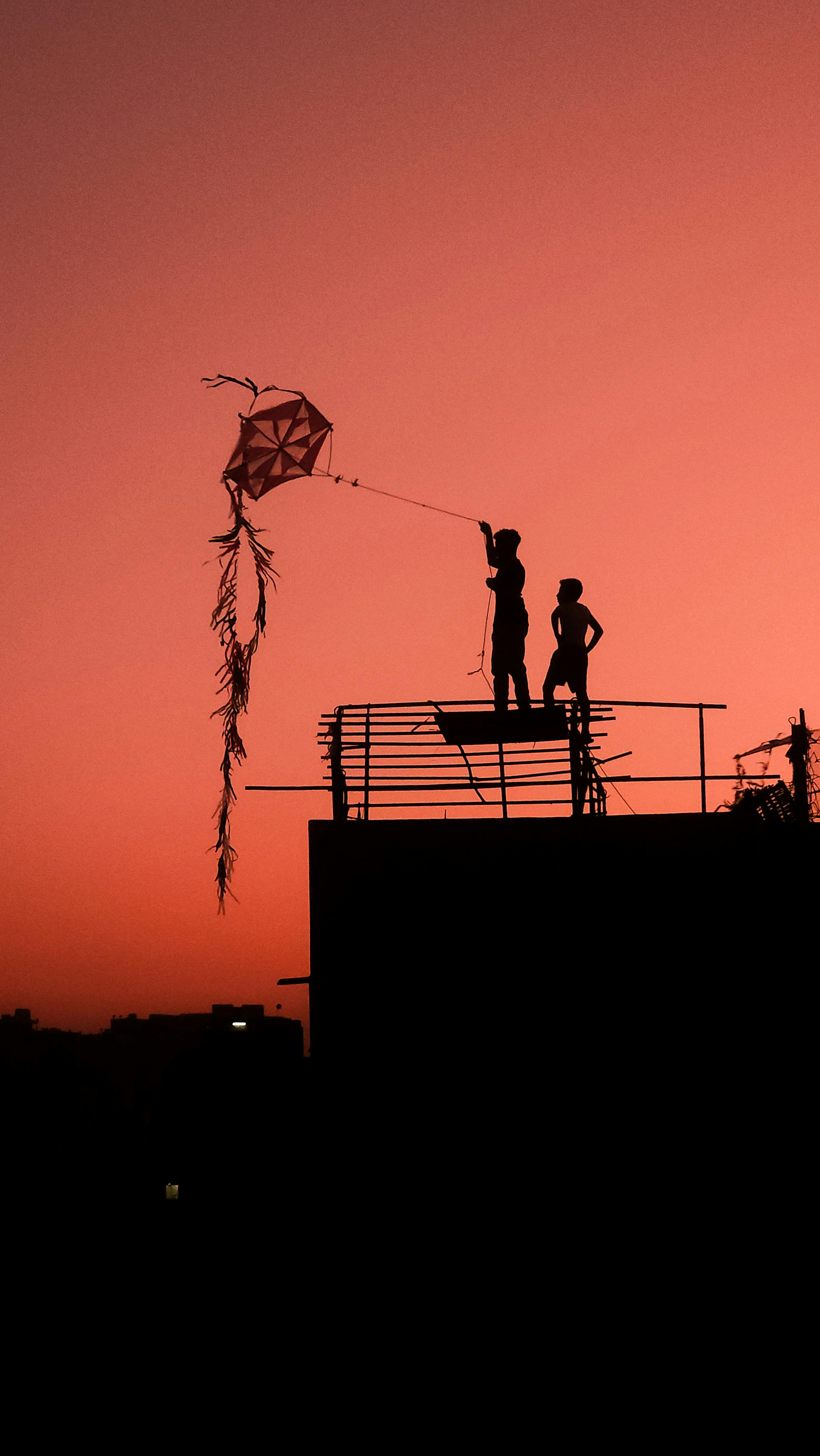 免費 夕陽西下，落日餘暉映照著屋頂上放風箏的兩個人的剪影。 圖庫相片