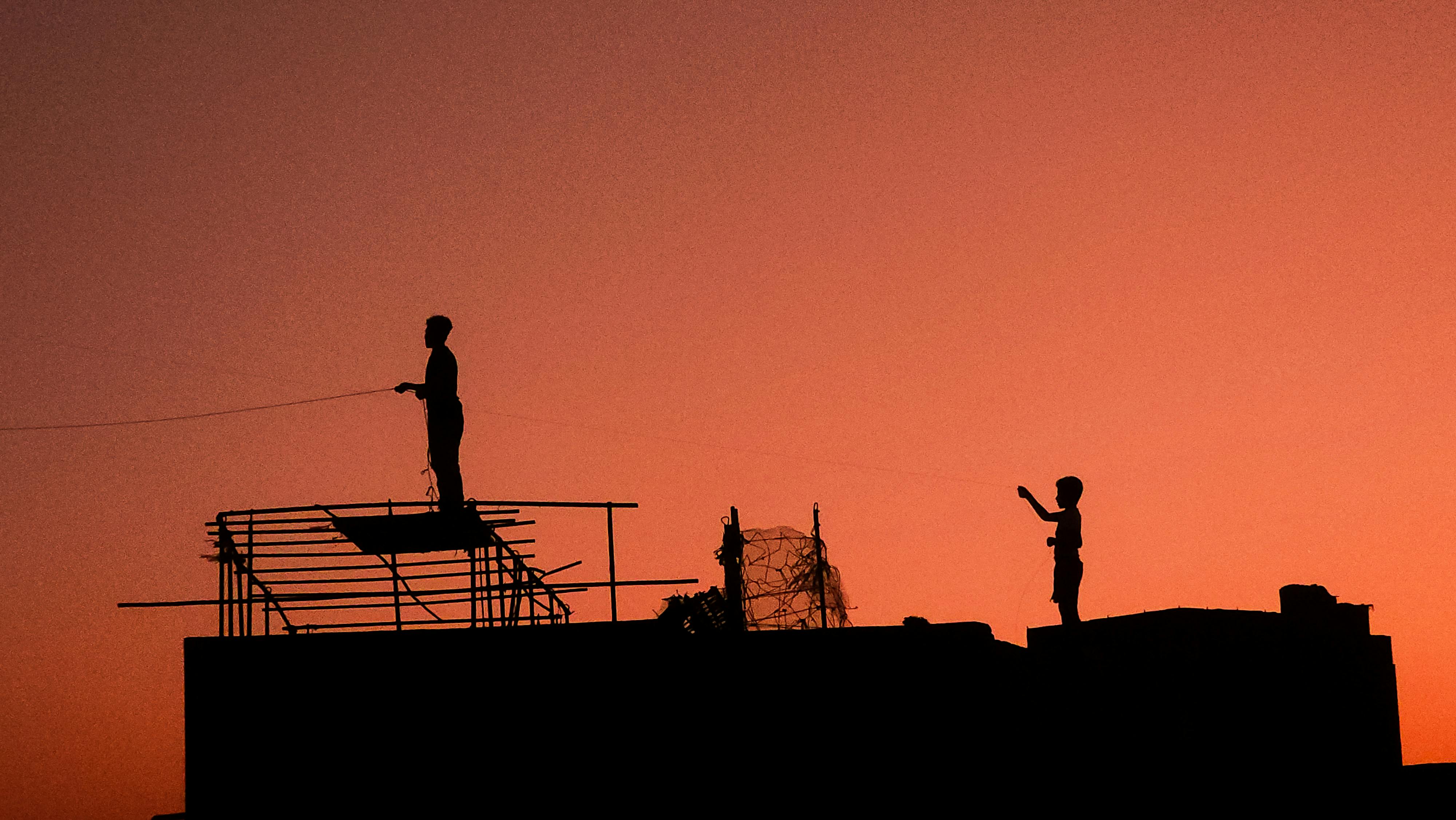 bezplatná Silueta dvou lidí pouštějících draky na pozadí zářivě oranžové oblohy při západu slunce. Zachycuje podstatu volného času a svobody. Základová fotografie