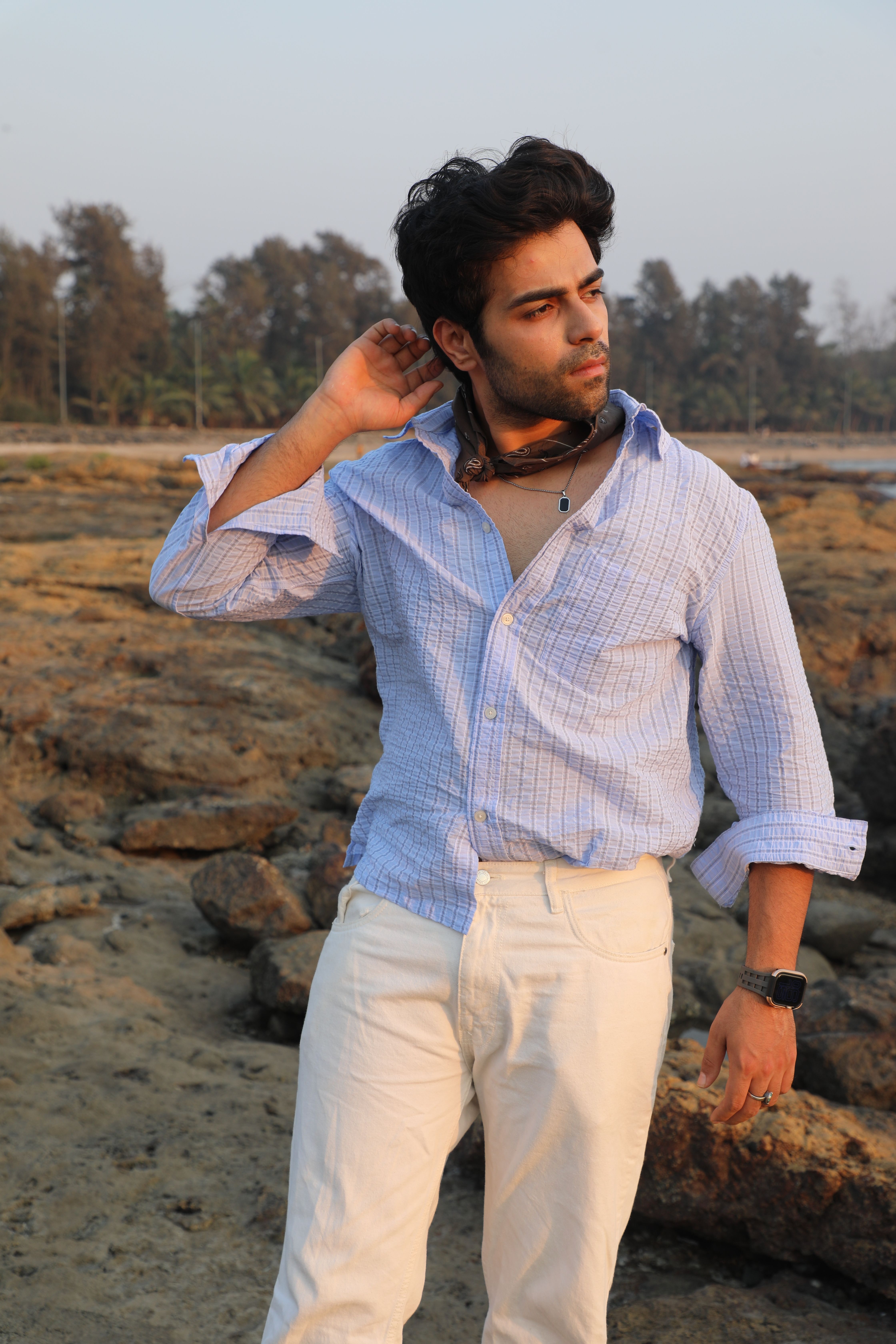 Young man in casual attire posing on a rocky beach during sunset.
