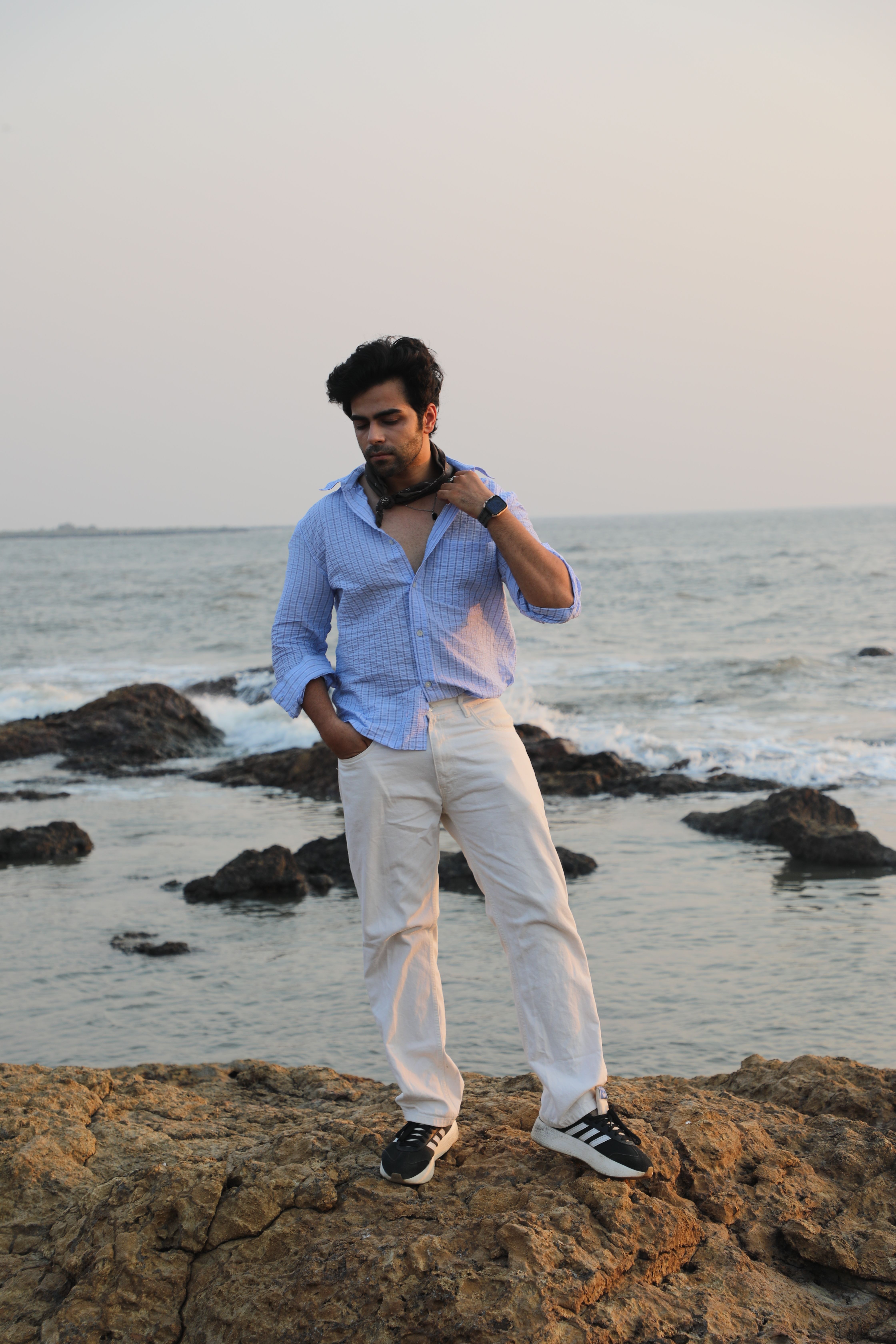 A stylish man poses on a rocky shore with an ocean backdrop during sunset.