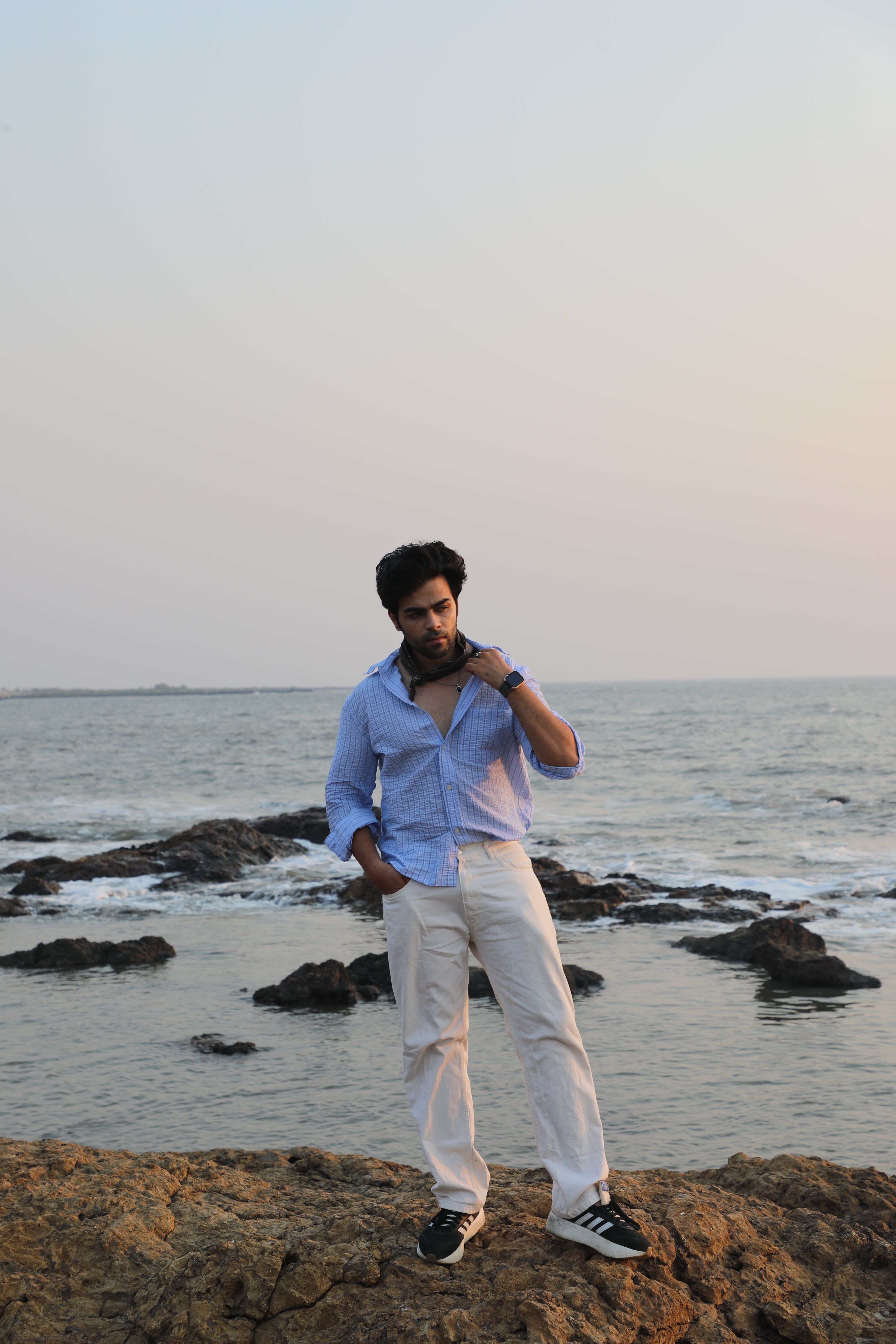 A man in casual attire poses on a rocky beach at sunset, with ocean waves in the background.