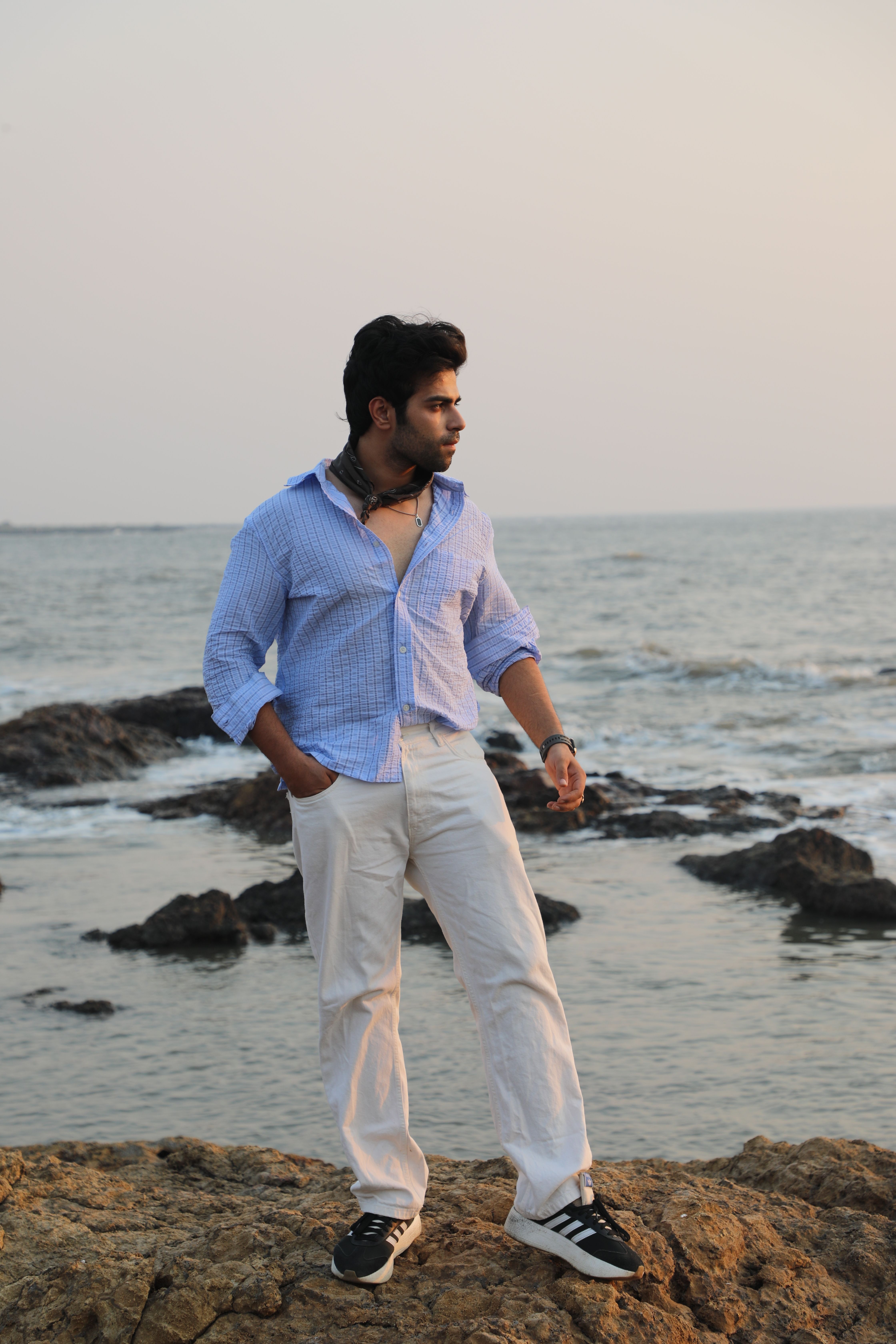 Young man standing on a rocky beach during sunset with ocean waves in the background, exuding a relaxed vibe.