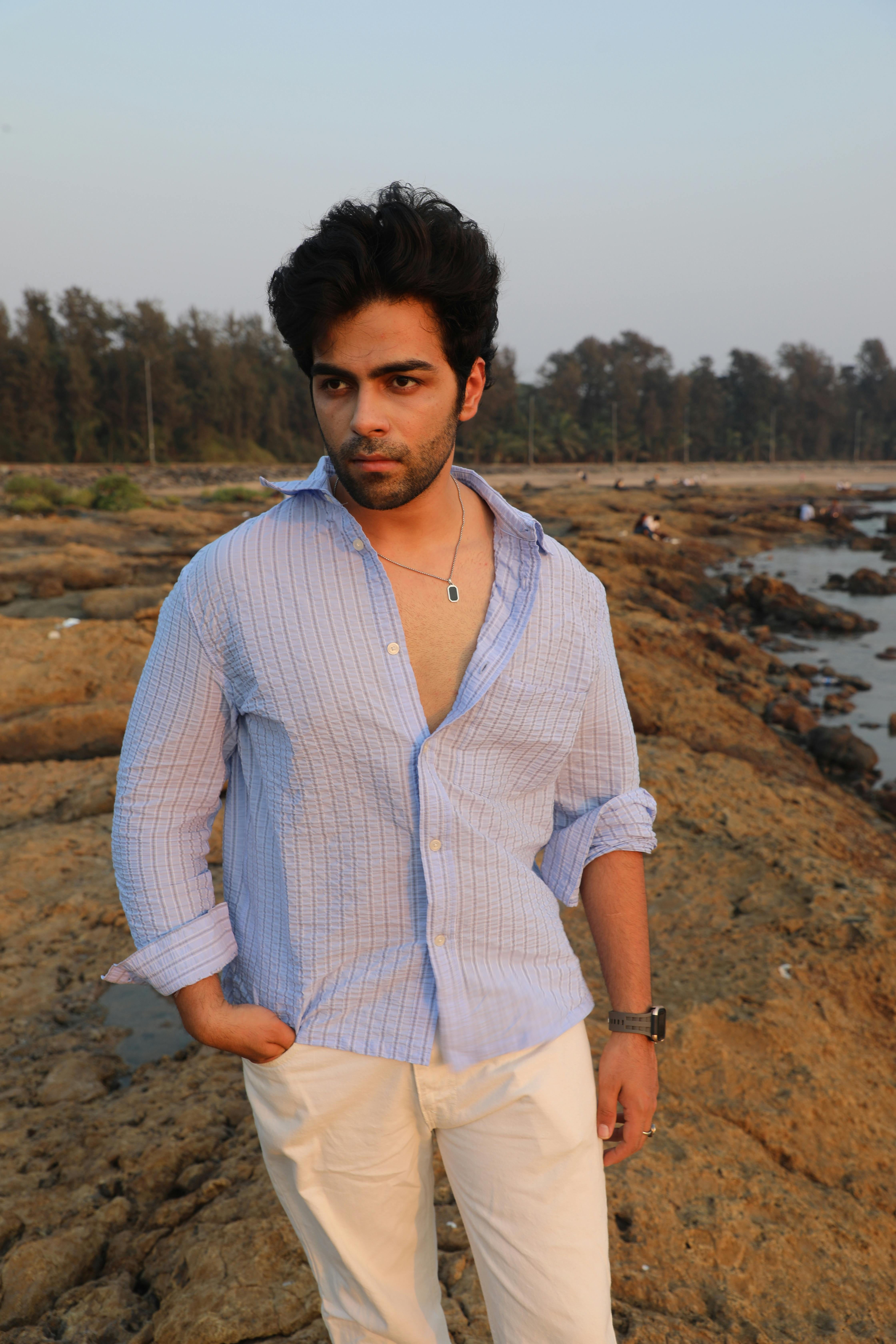 Stylish young adult man in blue shirt standing on a rocky coastline at sunset.