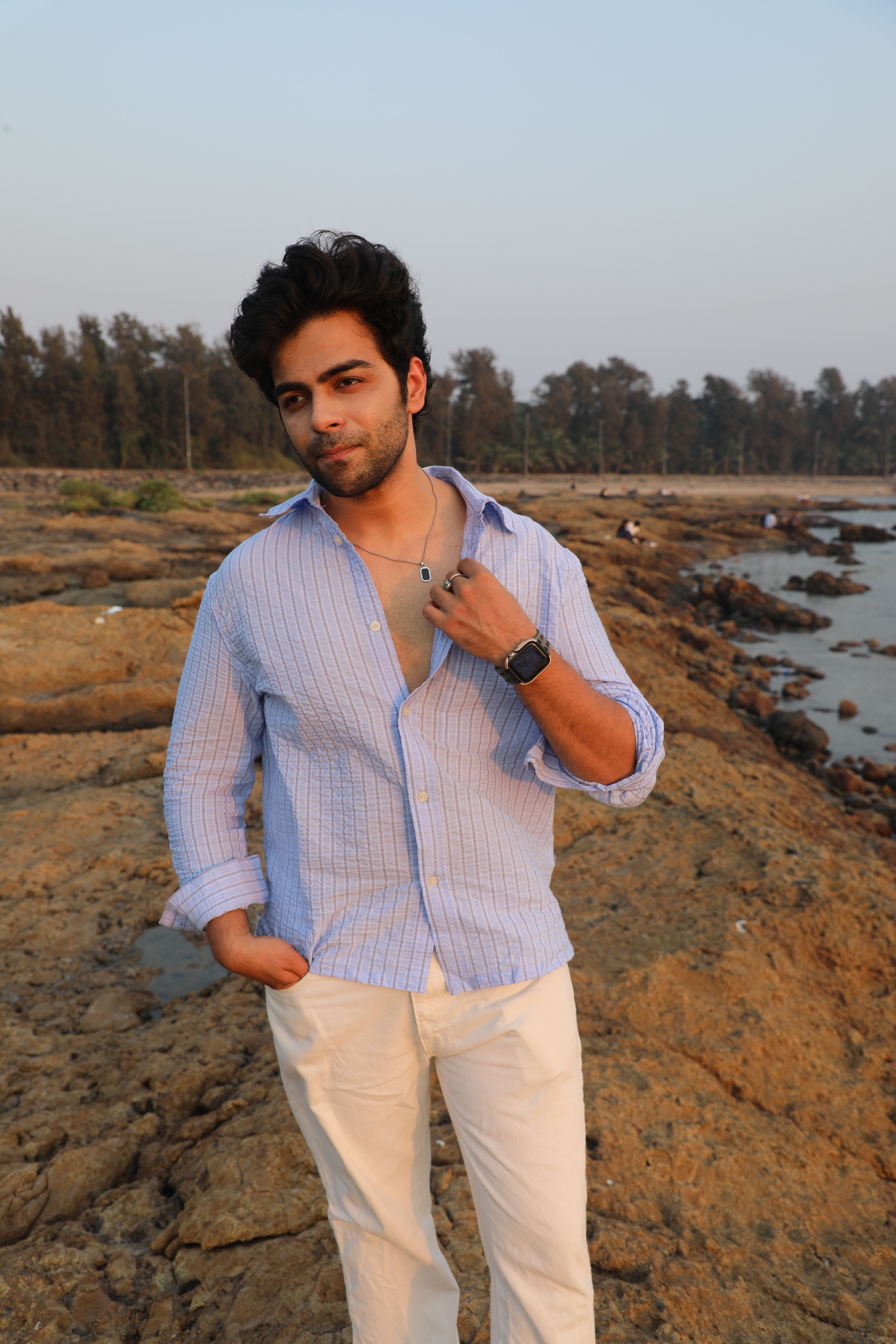 Adult man poses on a rocky beach during sunset, wearing a casual shirt and trousers.
