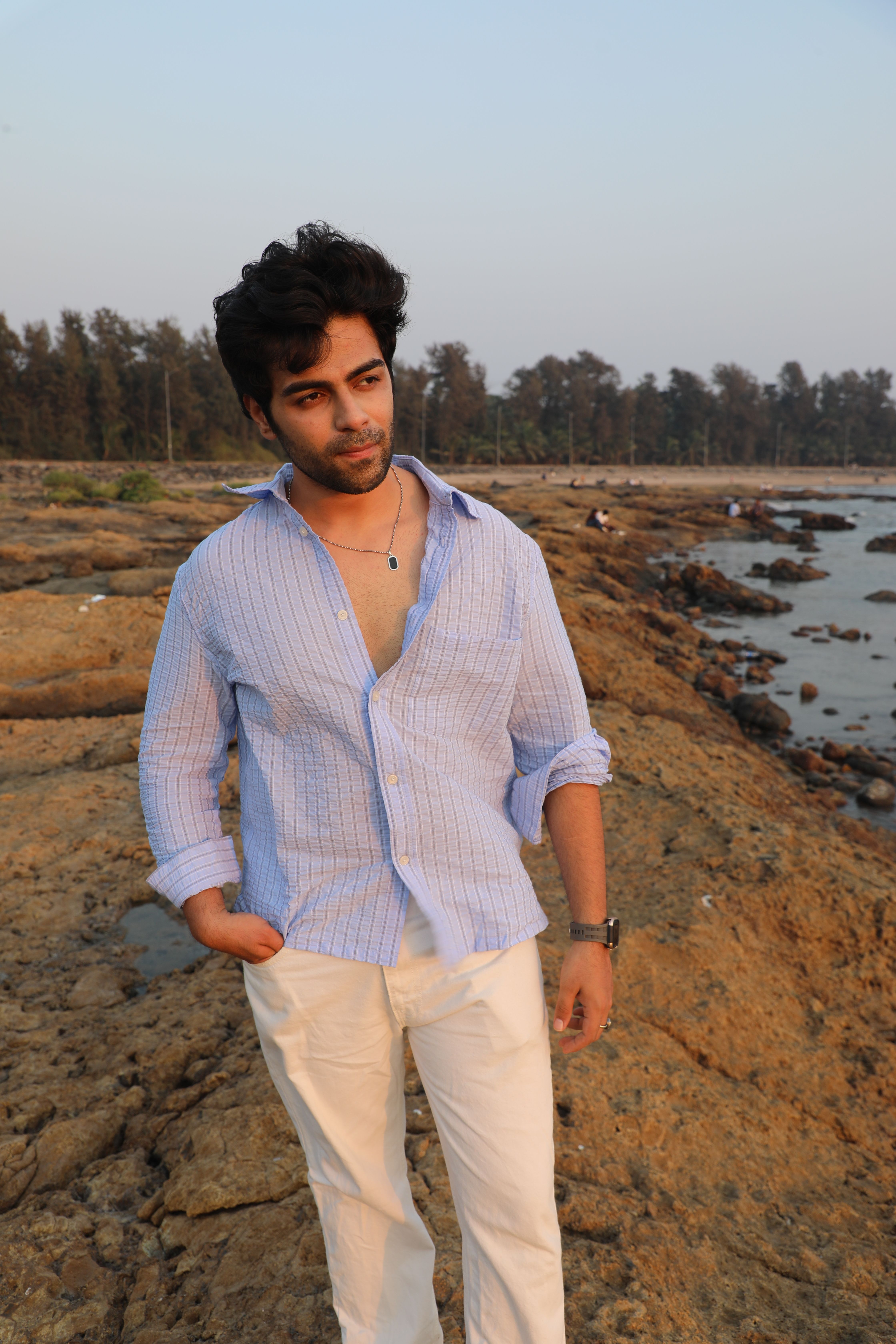 A young man in a casual shirt walking along a scenic rocky coastline during sunset.