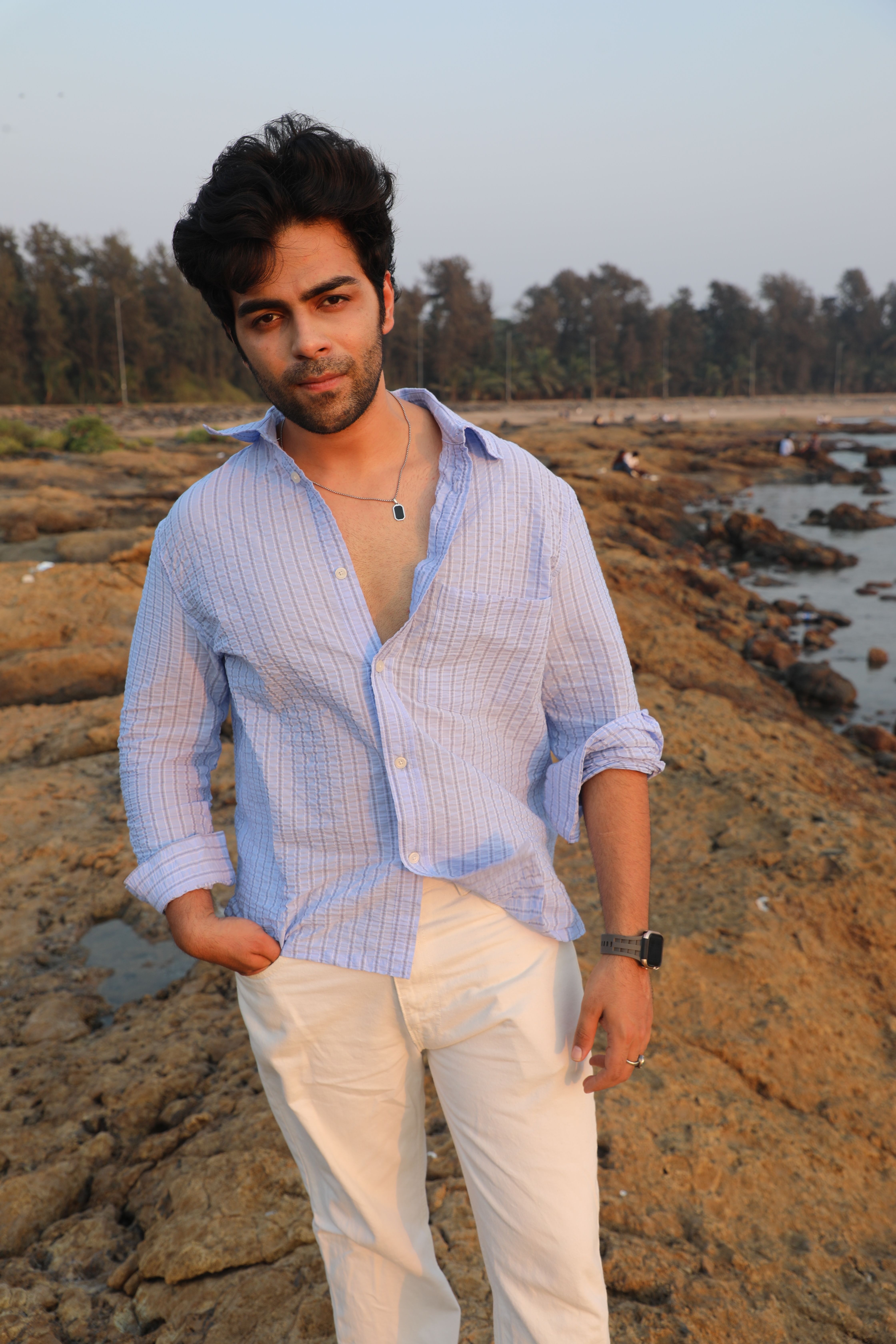 Young man stands confidently on a rocky beach during sunset, wearing casual attire.