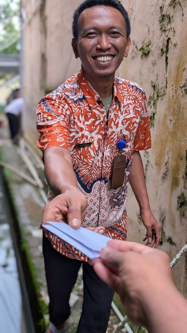 Smiling man in a colorful shirt hands an item in a narrow alleyway.