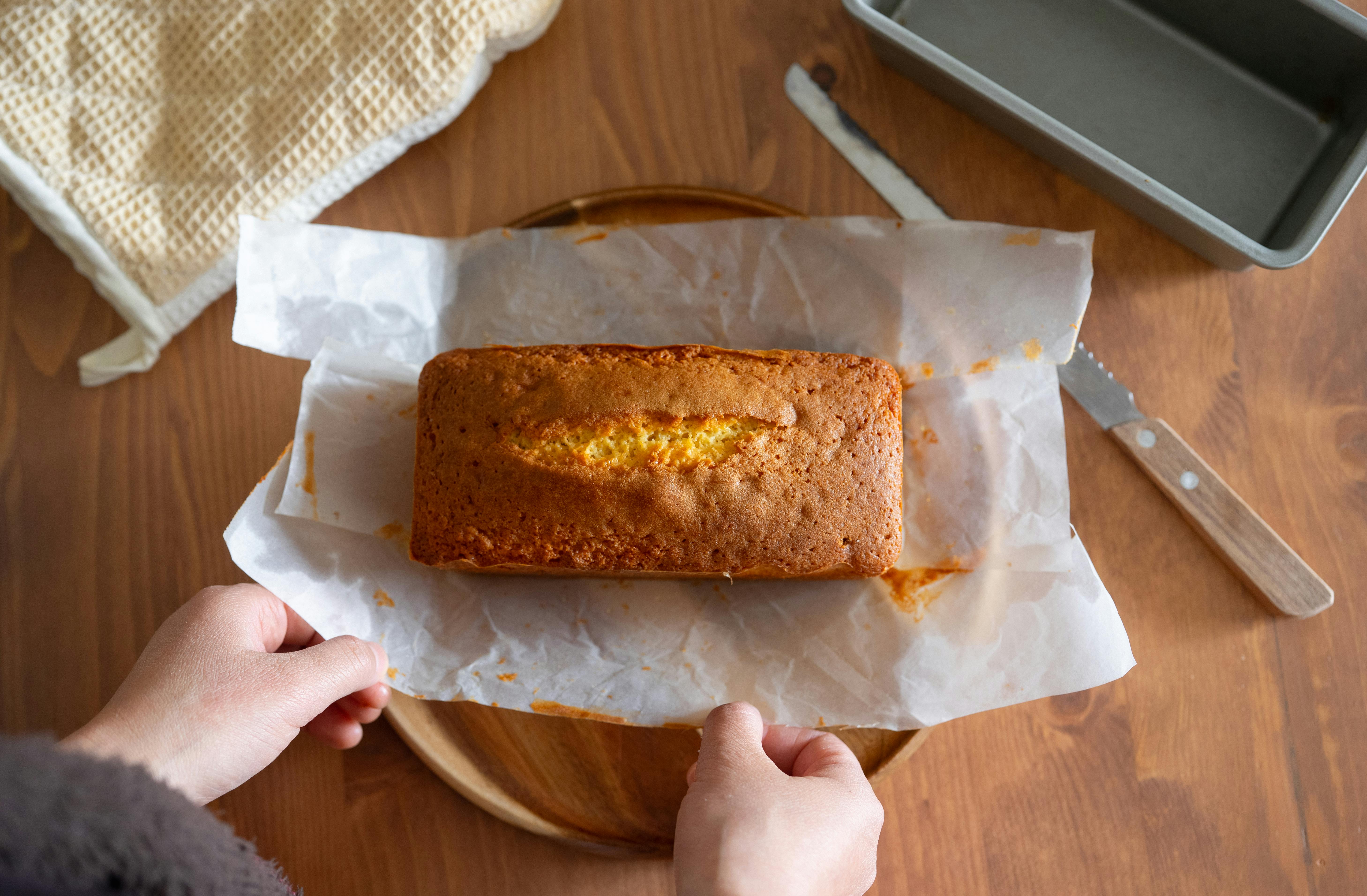 A freshly baked loaf cake on parchment paper being lifted on a wooden table. Perfect homemade baking scene.