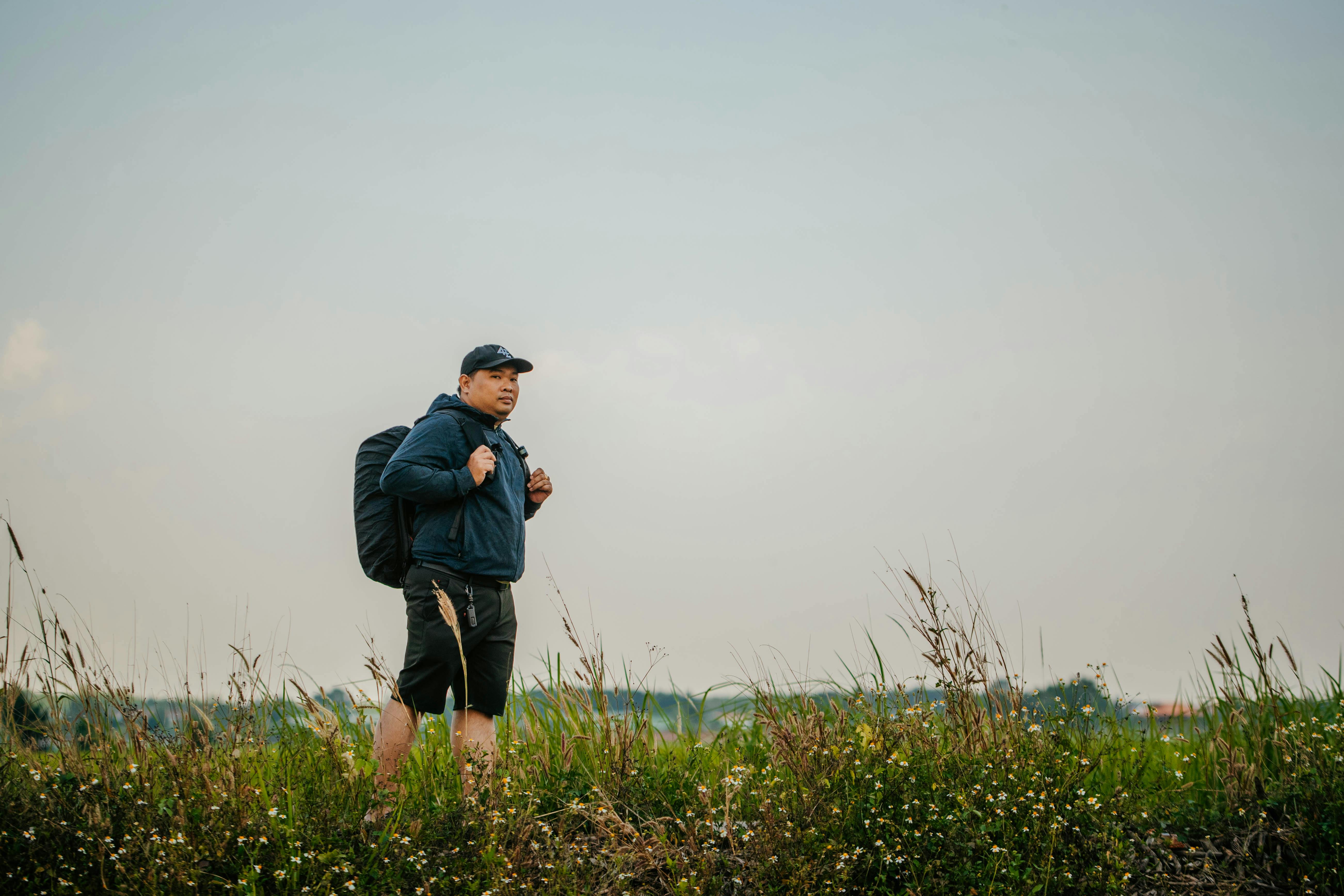 ingyenes Egy hétköznapi öltözéket viselő férfi hátizsákkal túrázik egy füves mezőn, élvezve a természetet. Stockfotó