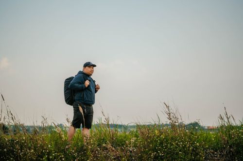 Free A man dressed in casual attire hiking through a grassy field with a backpack, enjoying nature. Stock Photo