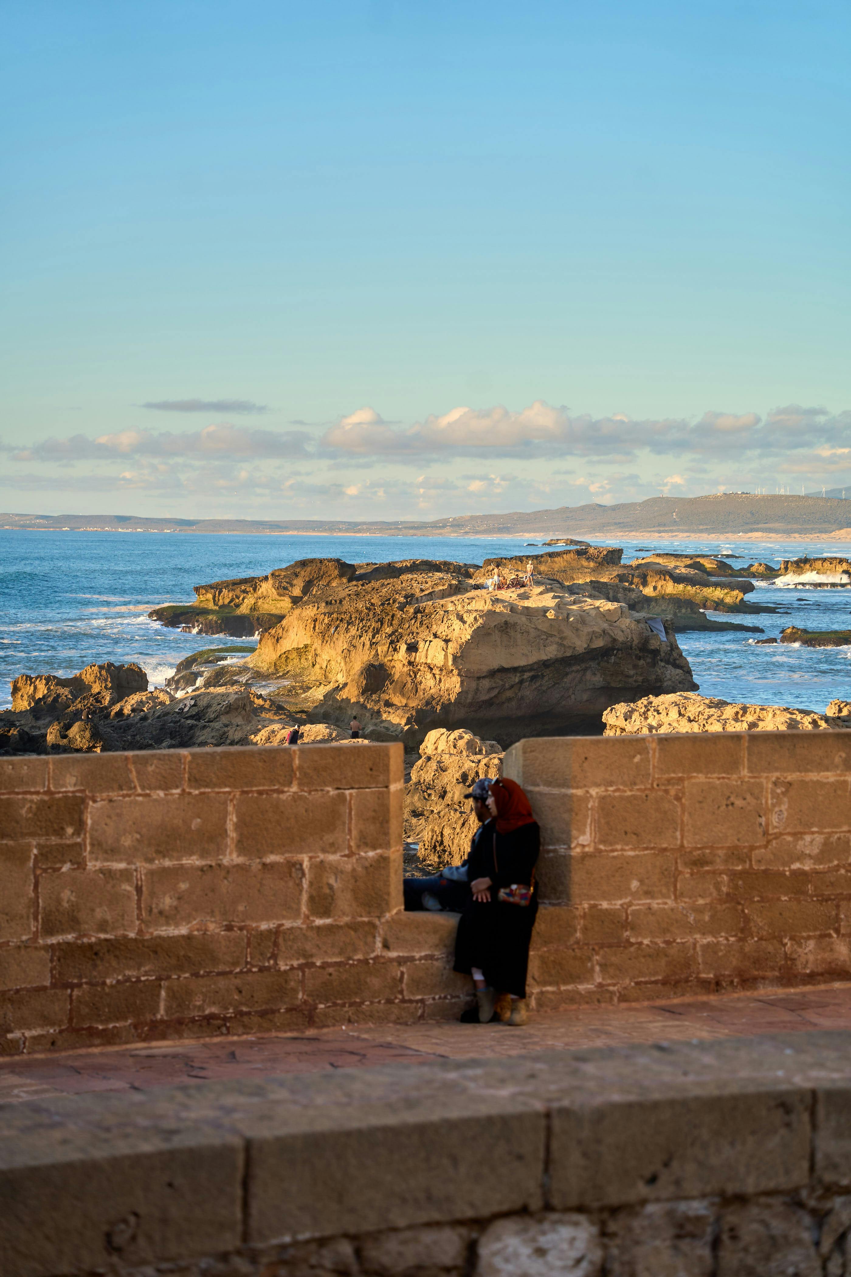 Gratis Una mujer vestida con atuendo tradicional disfruta de las vistas panorámicas del océano y la costa rocosa en Essaouira durante la puesta de sol. Foto de stock