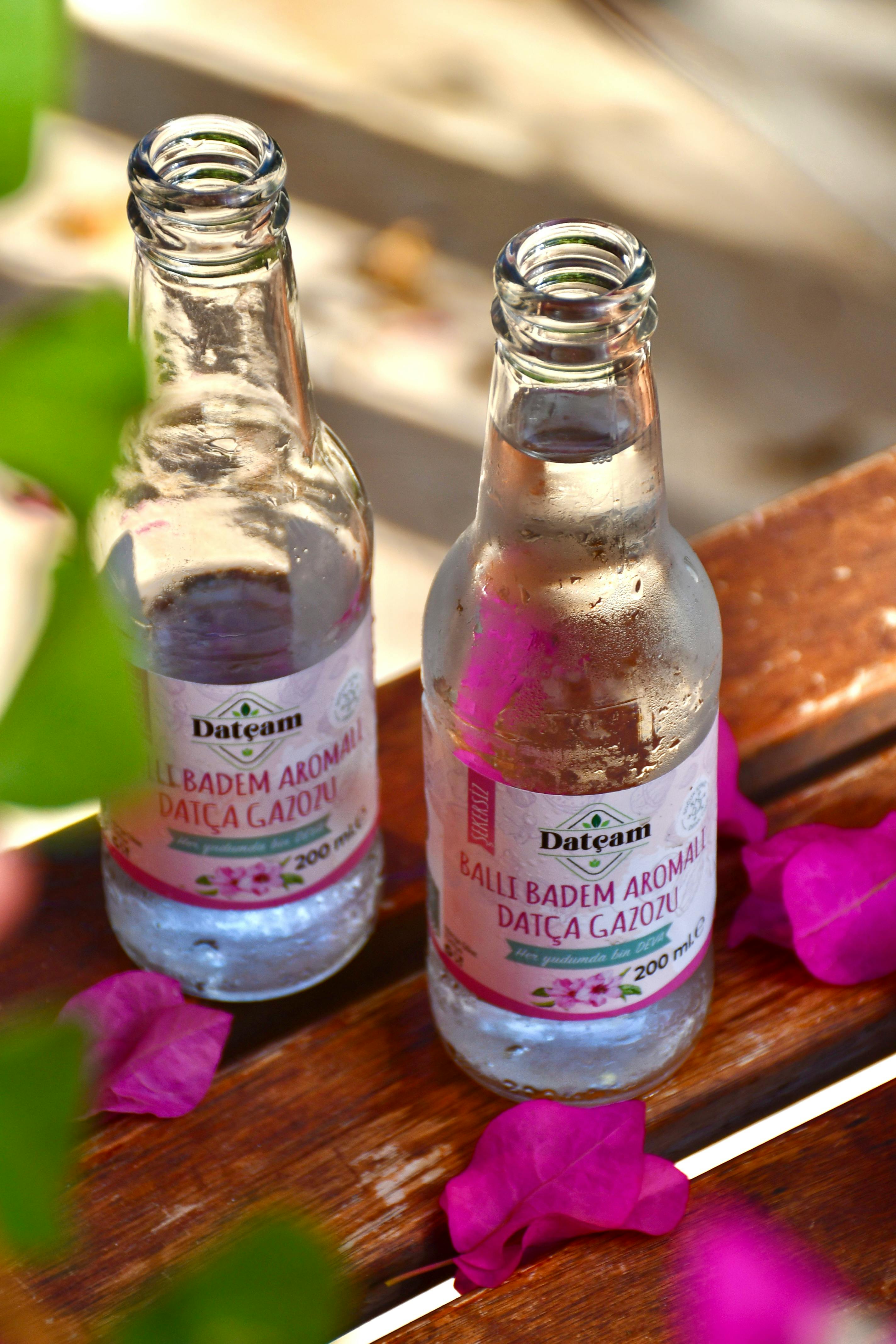 gratis Close-up van twee open flesjes amandelsoda met paarse bloemblaadjes op een houten tafel, die een verfrissende sfeer creëren. Stockfoto