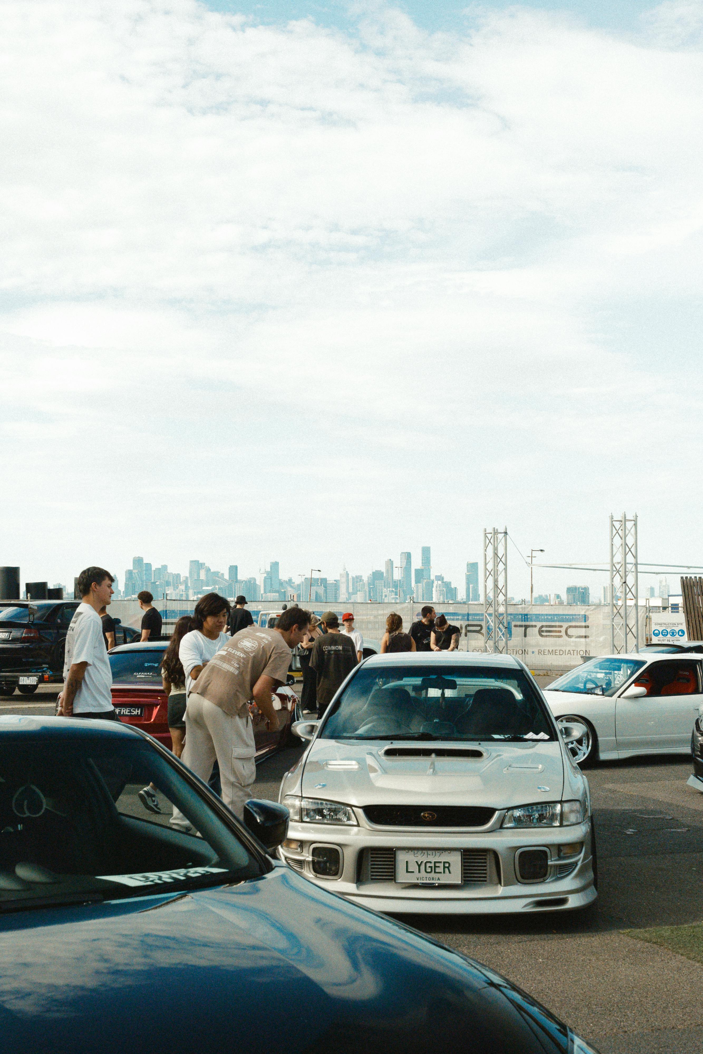Gratuit Un groupe de personnes rassemblées autour de voitures de sport lors d'un rassemblement automobile urbain ensoleillé avec vue sur la ville. Photos