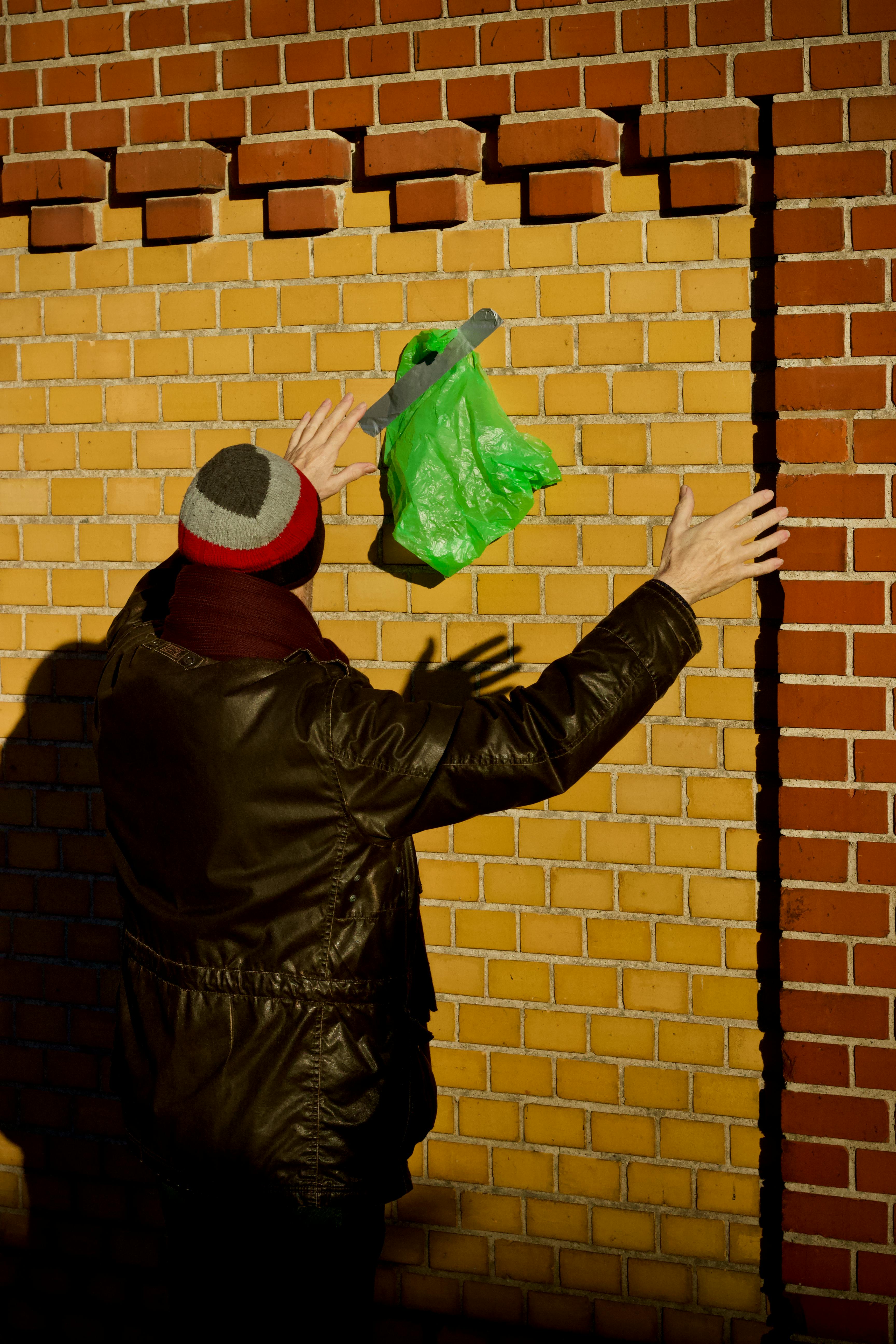 bezplatná Jednotlivec v teplém oblečení hodí za slunečného dne zelený igelitový sáček na cihlovou zeď. Základová fotografie