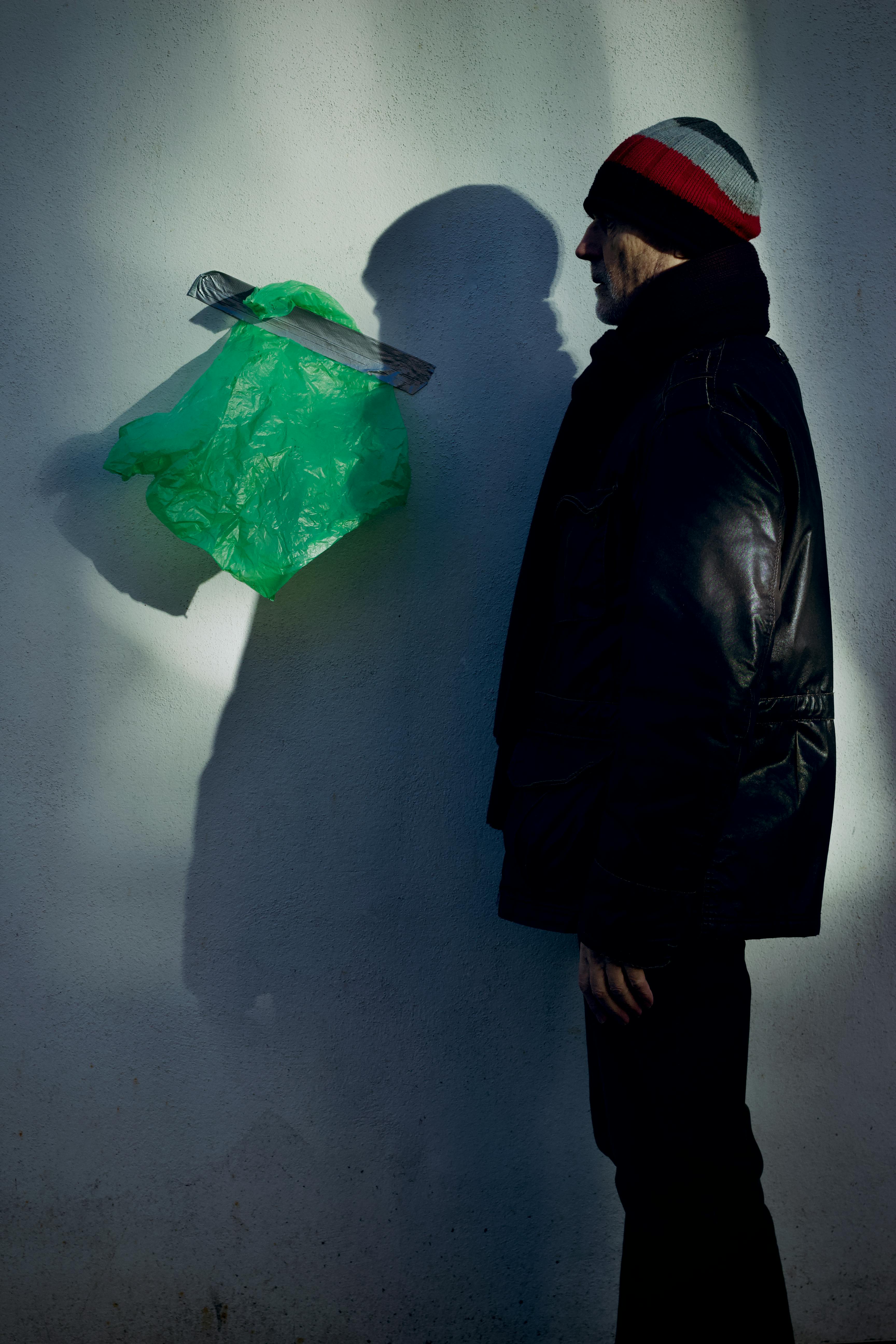 gratis Een man met een zwarte jas en muts observeert een drijvende groene plastic zak tegen een muur. Stockfoto