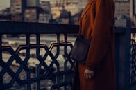一位穿着铁锈色外套的时尚女性站在城市桥上，展现着都市的时尚风格