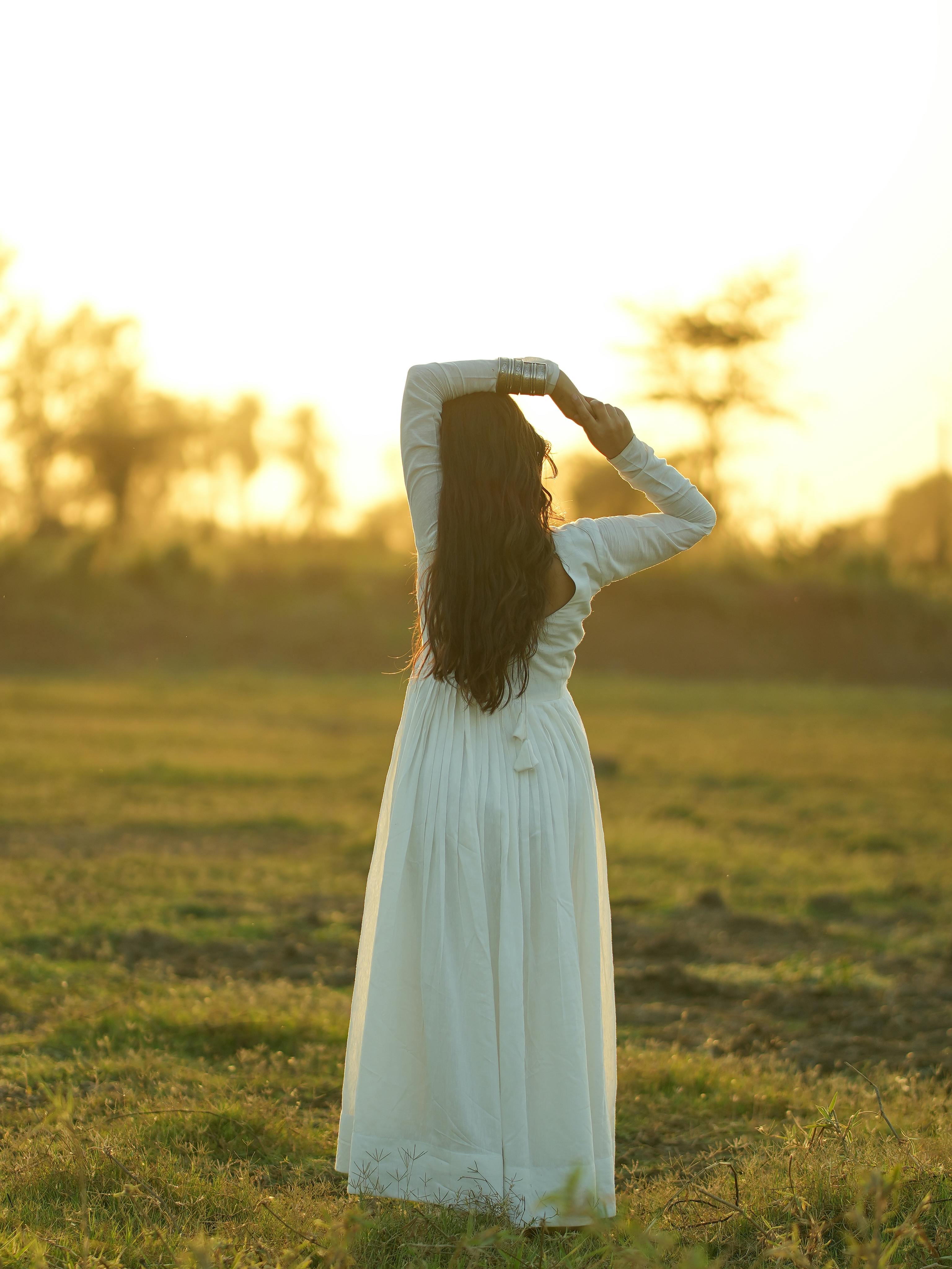 Zadarmo Žena v splývavých bielych šatách stojaca na poli pri západe slnka a objímajúca prírodu. Fotka z fotobanky