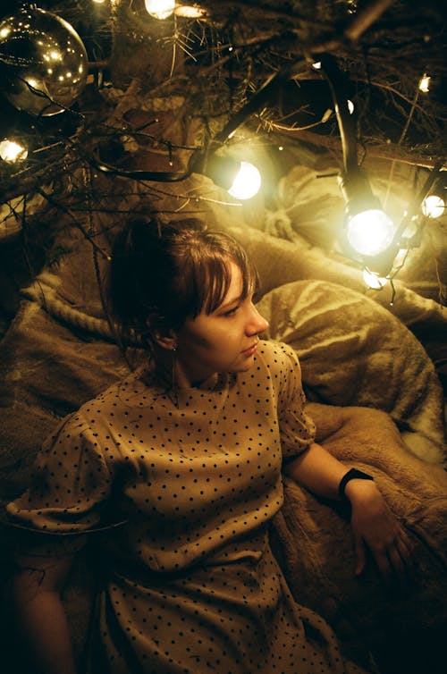 From above of dreamy young female in dress sitting alone under bright light of glowing lamps