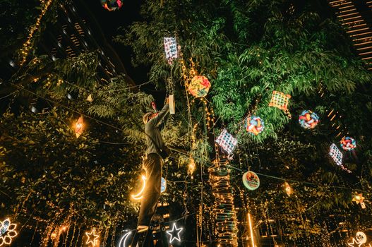 A person decorates a tree with vibrant lights and ornaments during a night festival.
