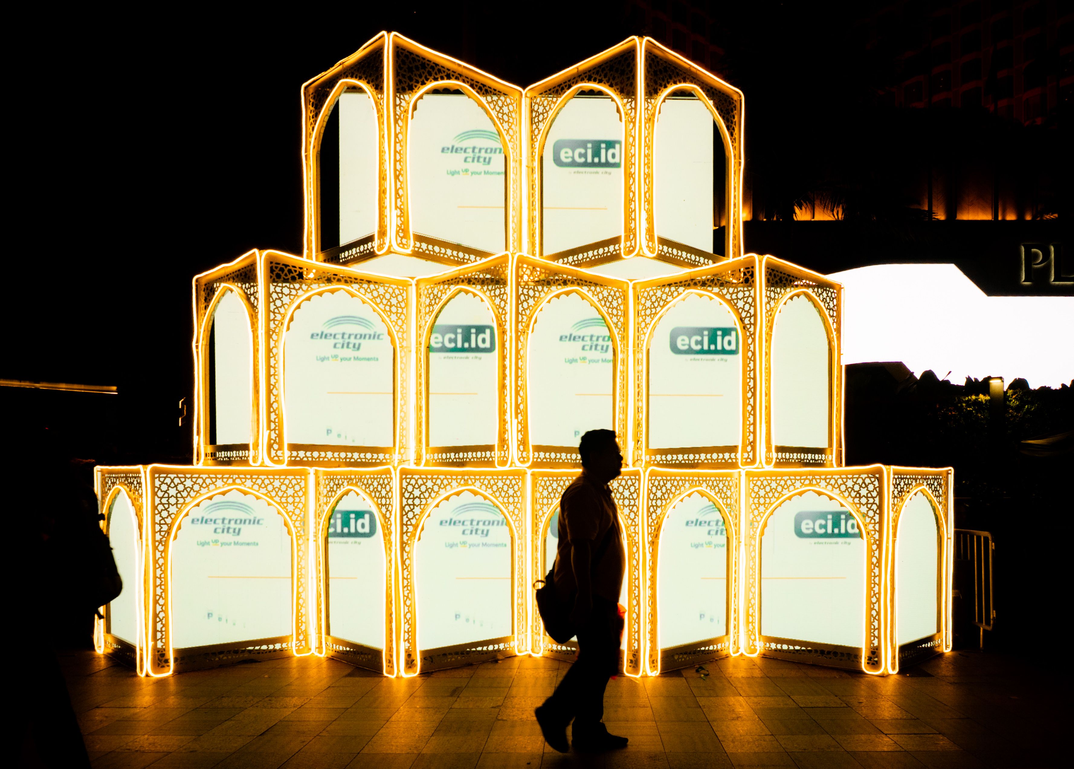 A striking backlit cube structure in a night market with silhouetted pedestrian.
