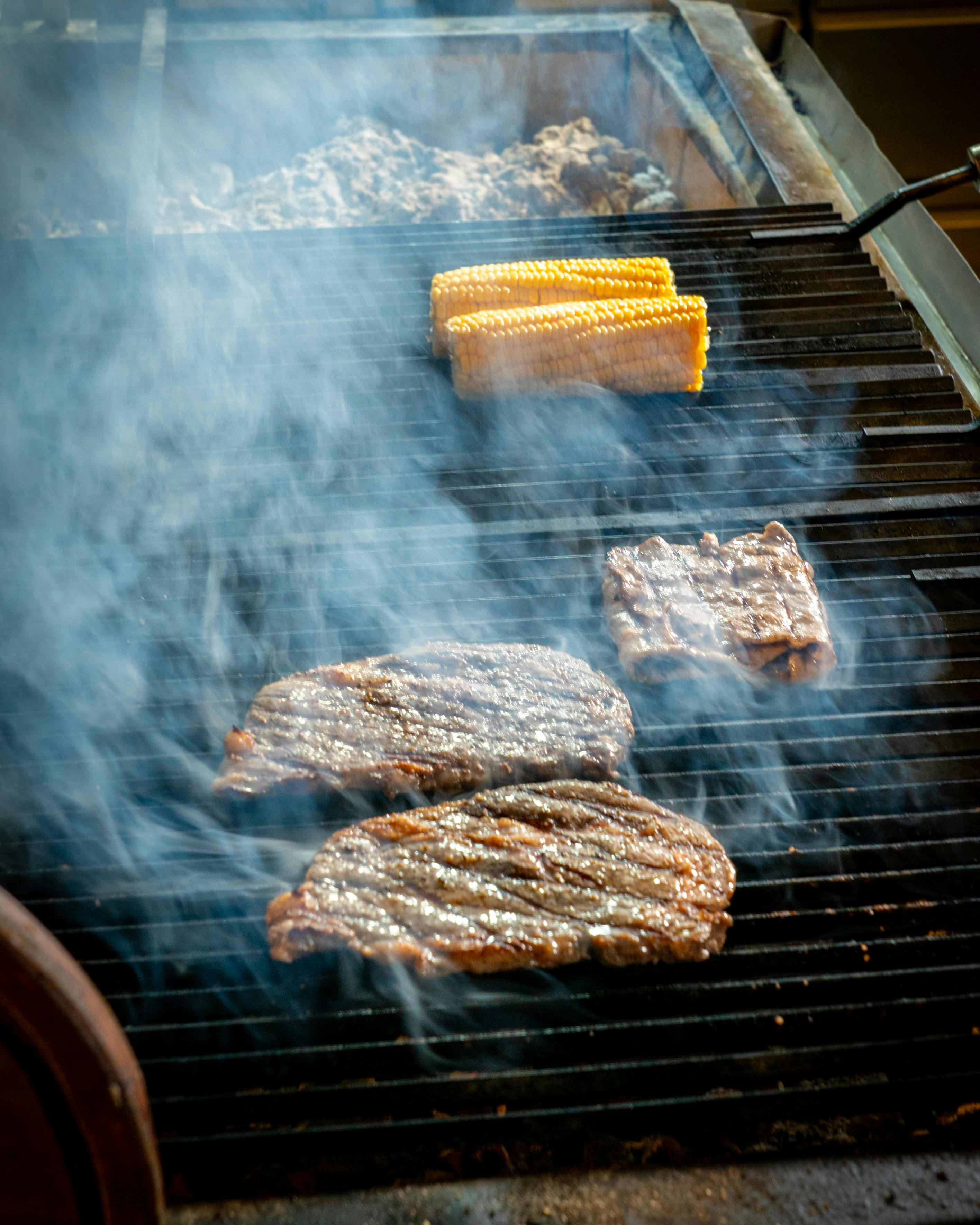 bezplatná Šťavnaté steaky a kukuřičný klas grilované s kouřem stoupajícím na barbecue. Základová fotografie