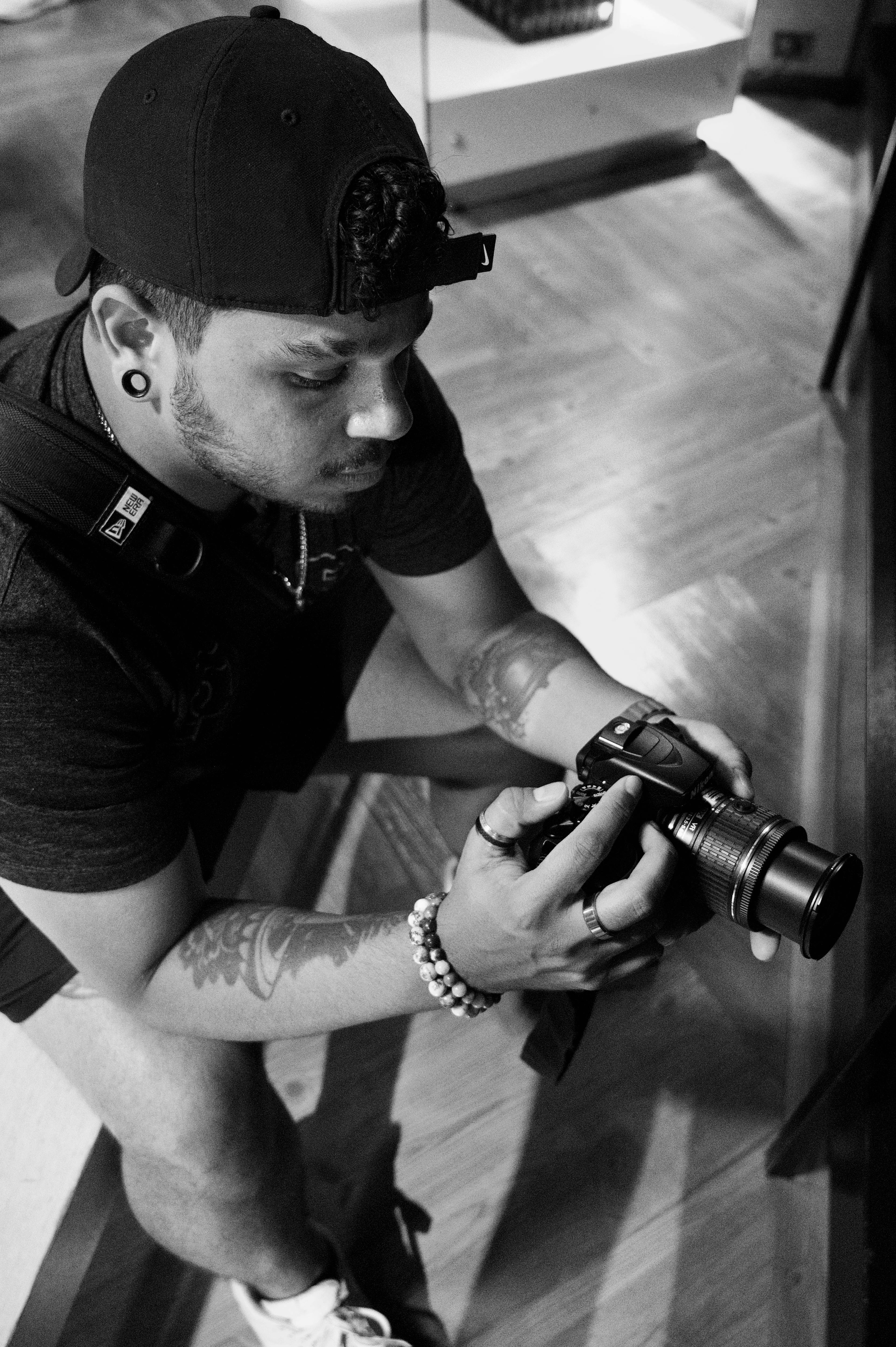 ฟรี ช่างภาพหนุ่มที่มีรอยสักและสวมหมวกกำลังตรวจสอบการตั้งค่ากล้องภายในอาคาร คลังภาพถ่าย