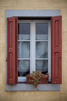 A picturesque Italian window with red shutters and a potted plant in Venzone, Italy.