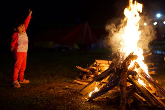 A young child in pajamas raises a hand excitedly beside a glowing campfire under the night sky.