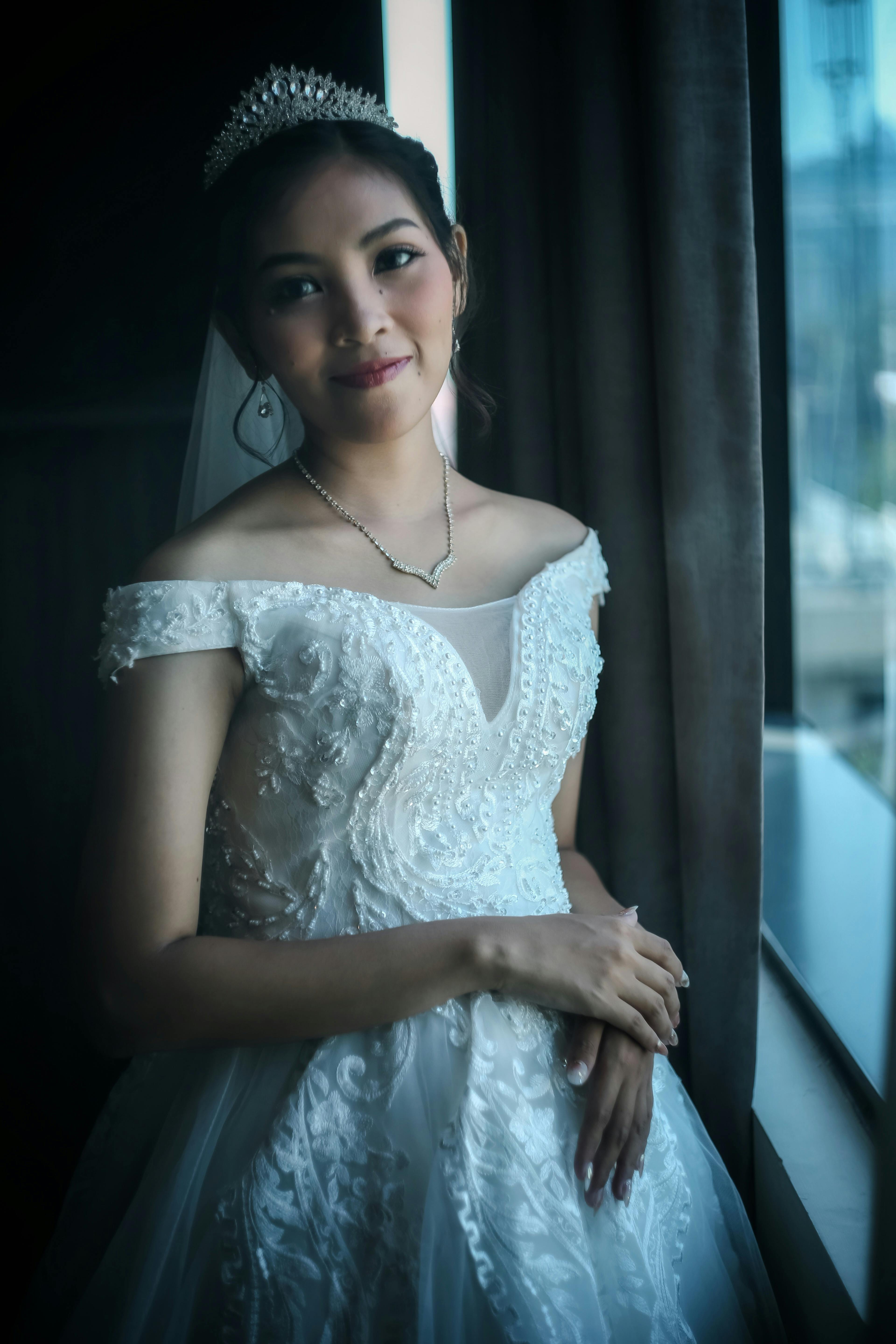 Kostnadsfria En vacker brud i vit brudklänning poserar elegant vid ett fönster och visar upp sitt fridfulla uttryck och sin brudklädsel. Stock foto