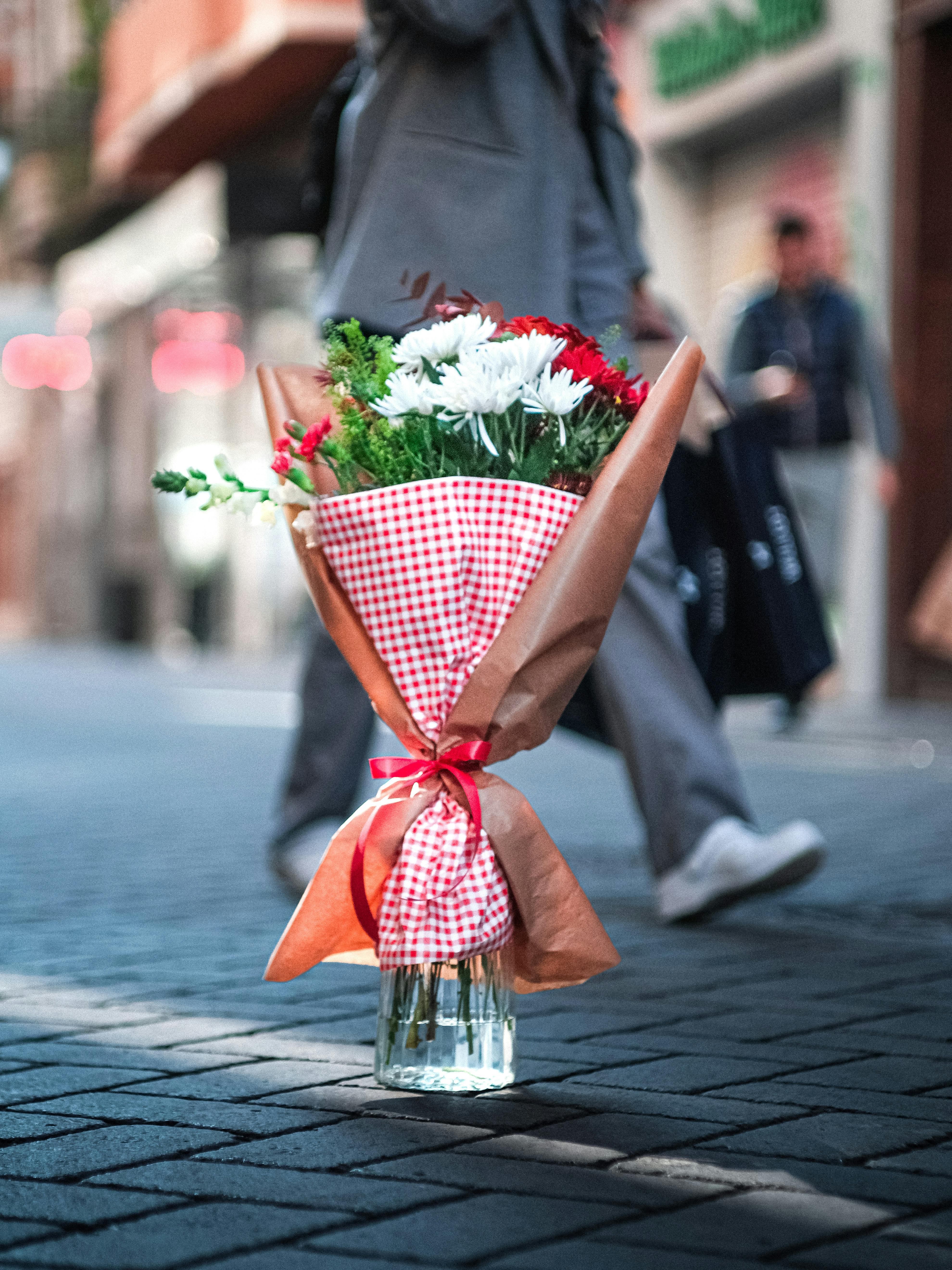 Miễn phí Một bó hoa rực rỡ được gói trong giấy kẻ caro đỏ đặt trên đường phố, phía sau là những người đi bộ mờ ảo. Ảnh lưu trữ