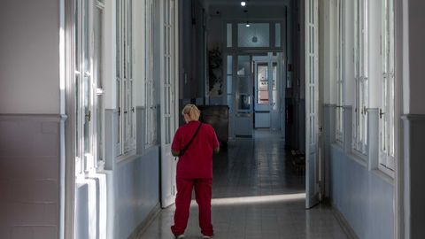 A nurse in red scrubs walks through a sunlit corridor in a hospital setting.