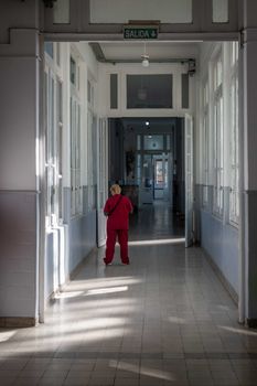 A nurse in red scrubs walks through a sunlit corridor in a hospital setting.