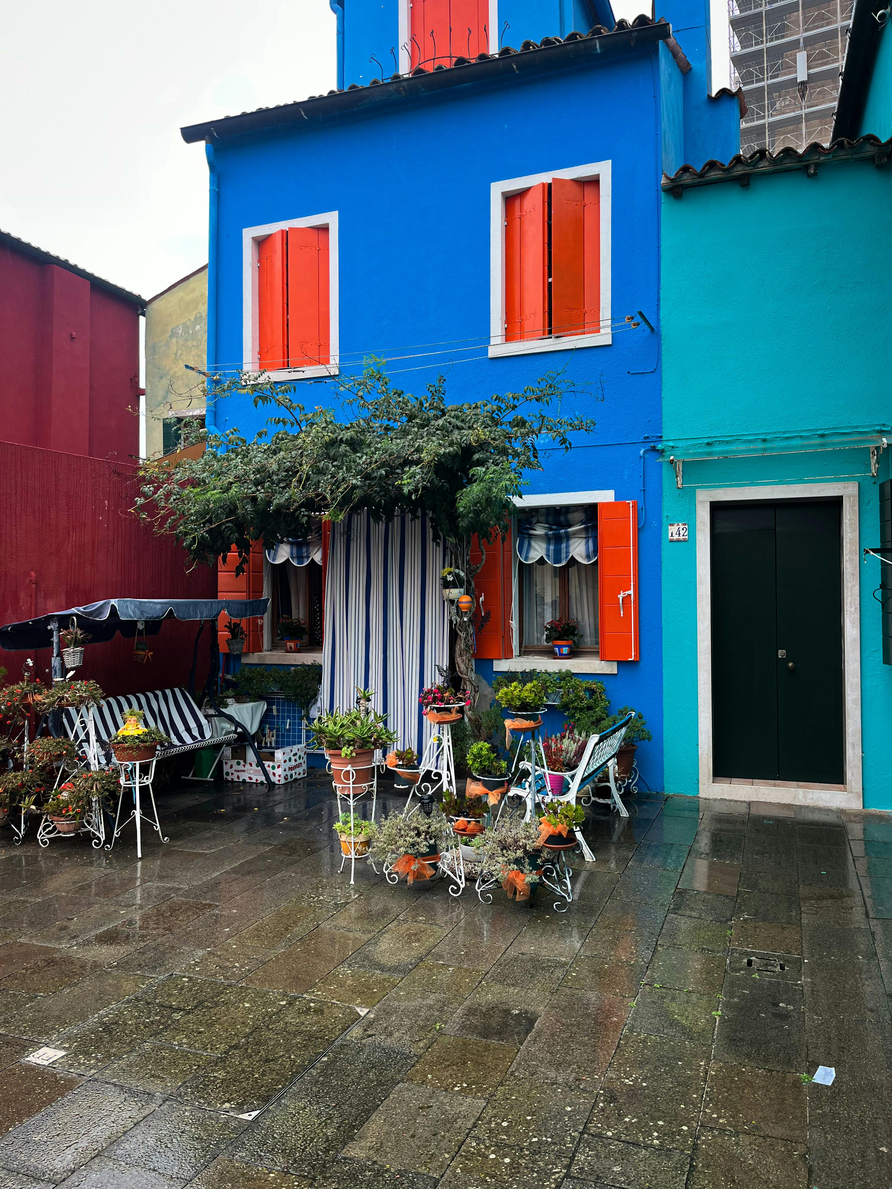免费 意大利布拉诺一条雨中的街道上，一栋充满活力的蓝色房子，周围环绕着植物和户外座椅。 素材图片