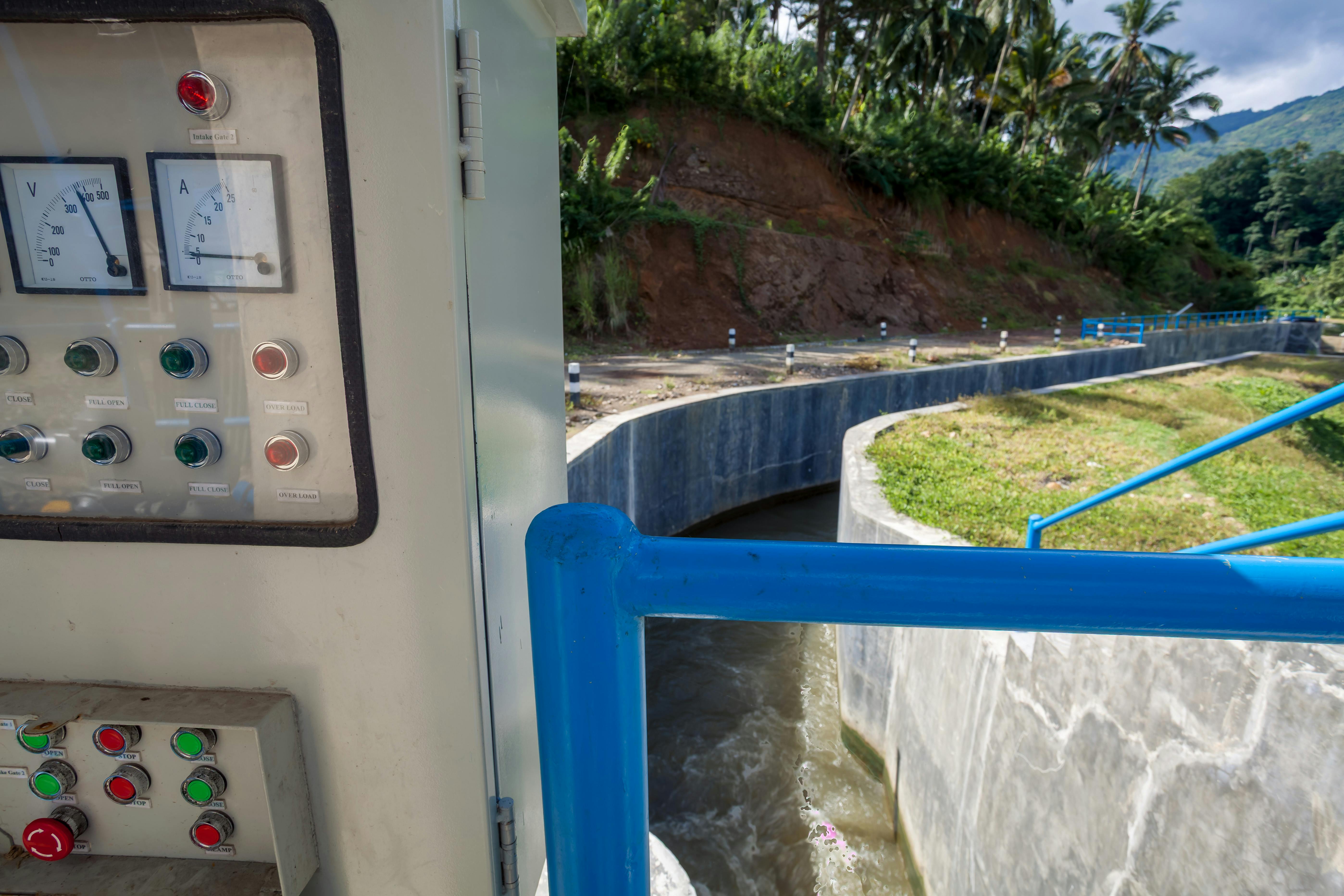 免費 印尼一條水渠附近的控制面板，背景是鬱鬱蔥蔥的綠植和起伏的山丘。 圖庫相片