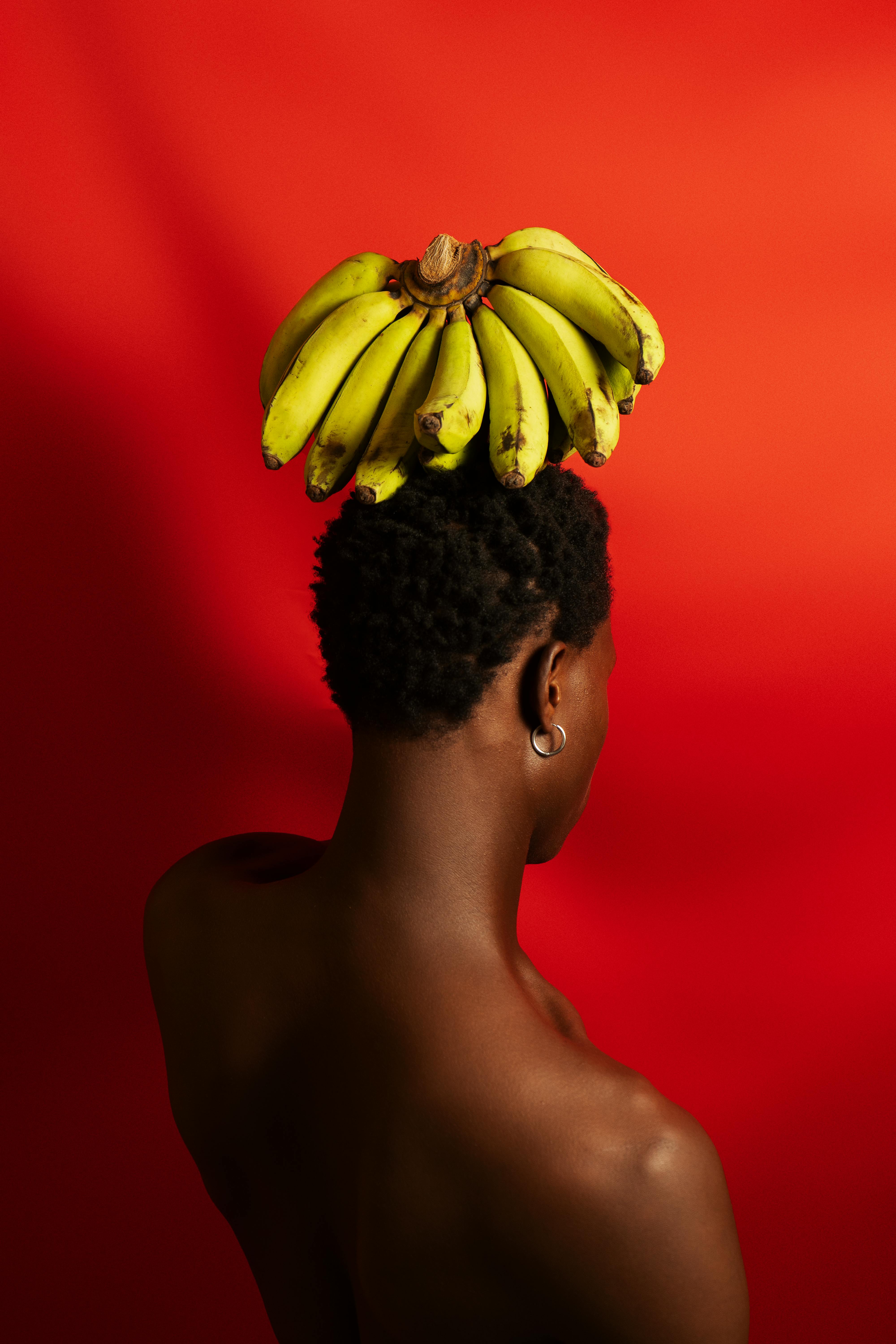 ฟรี ภาพถ่ายบุคคลเชิงสร้างสรรค์ของนางแบบชาวแอฟริกันที่มีกล้วยอยู่บนศีรษะ โดยมีฉากหลังเป็นสีแดง คลังภาพถ่าย