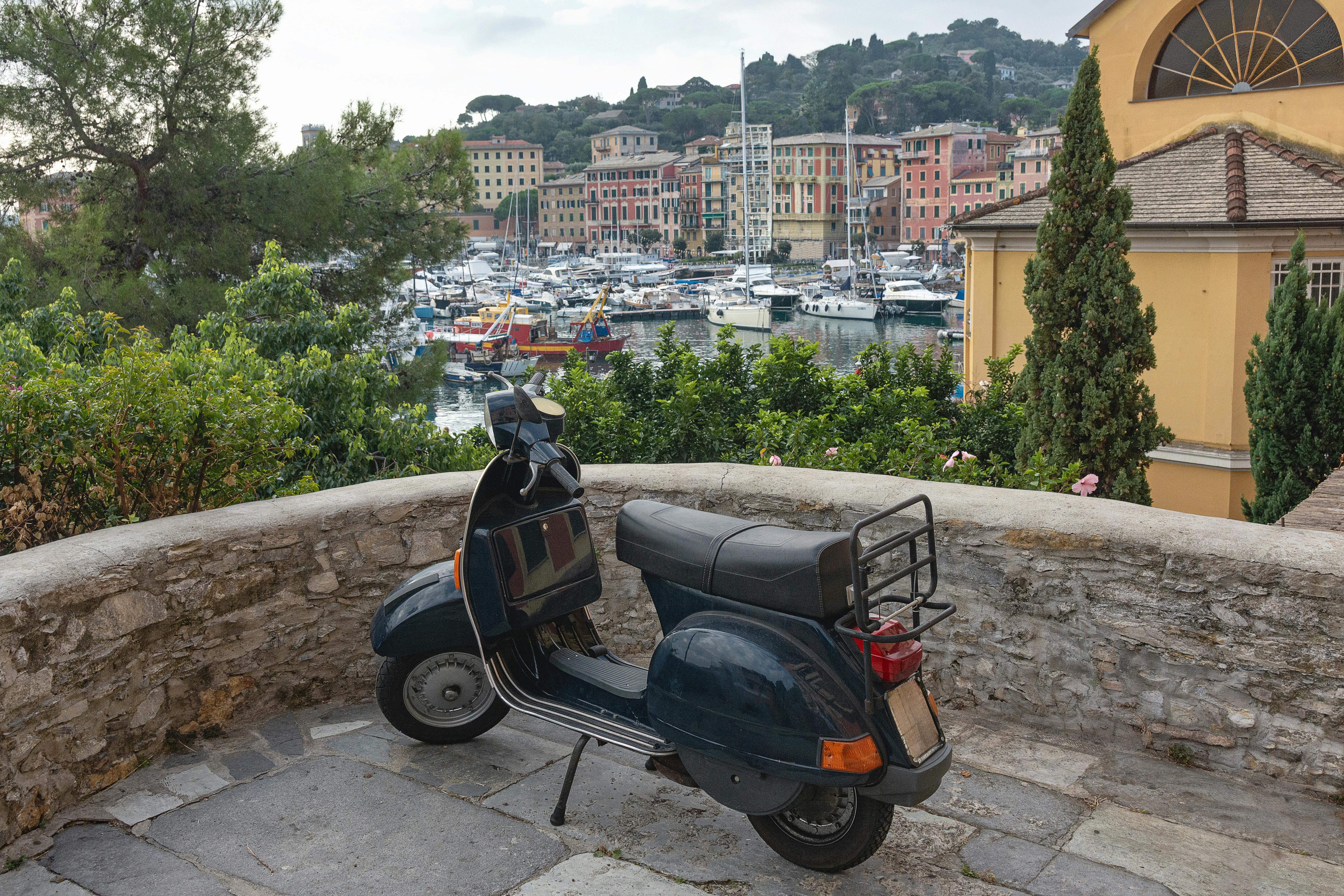 免費 一輛經典的 Vespa 摩托車停在義大利風景如畫的港口邊，周圍是色彩繽紛的建築和船隻。 圖庫相片