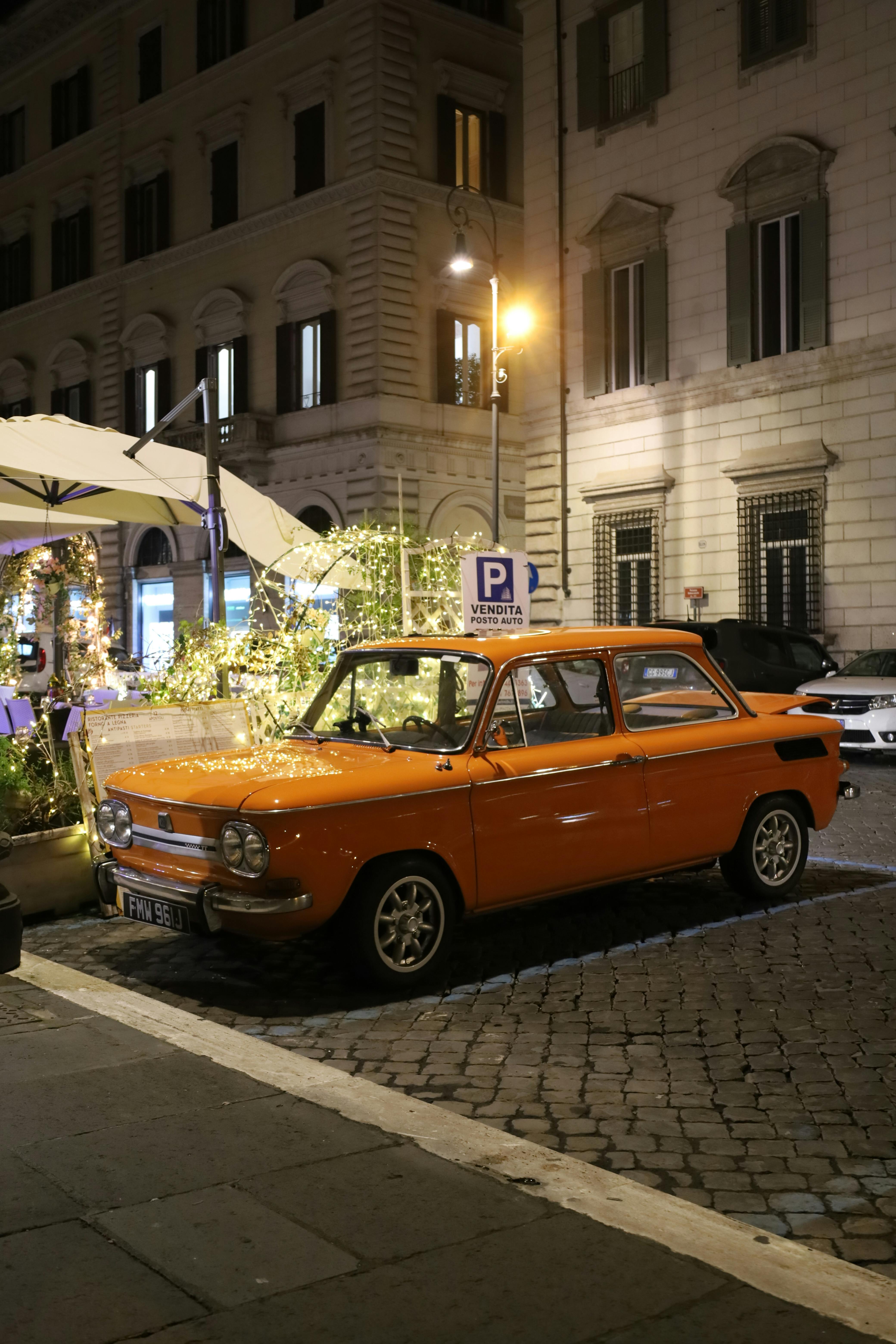 Miễn phí Một chiếc xe hơi màu cam cổ điển đậu trên con phố lát đá cuội ở Rome, Ý vào ban đêm, được chiếu sáng bởi đèn đường. Ảnh lưu trữ